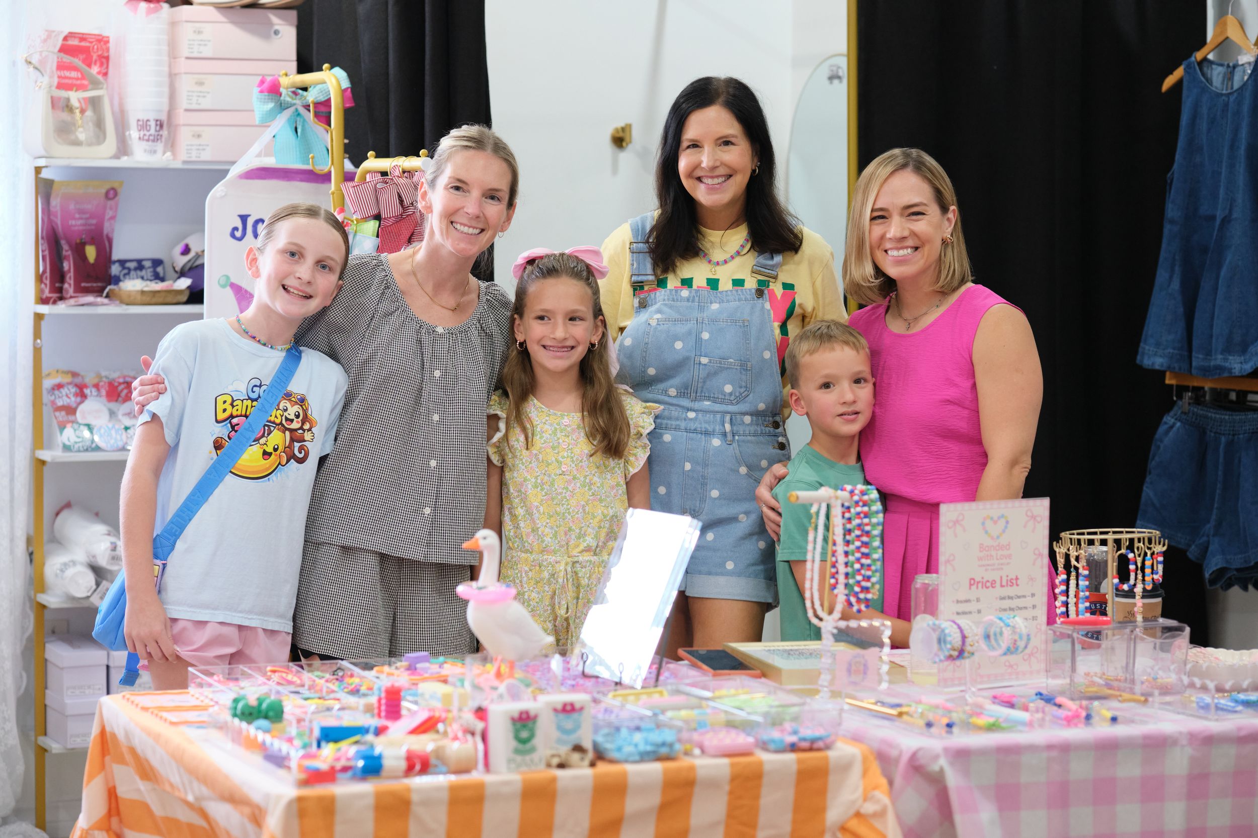 Two women and four children smile together at a colorful craft and jewelry vendor booth indoors.