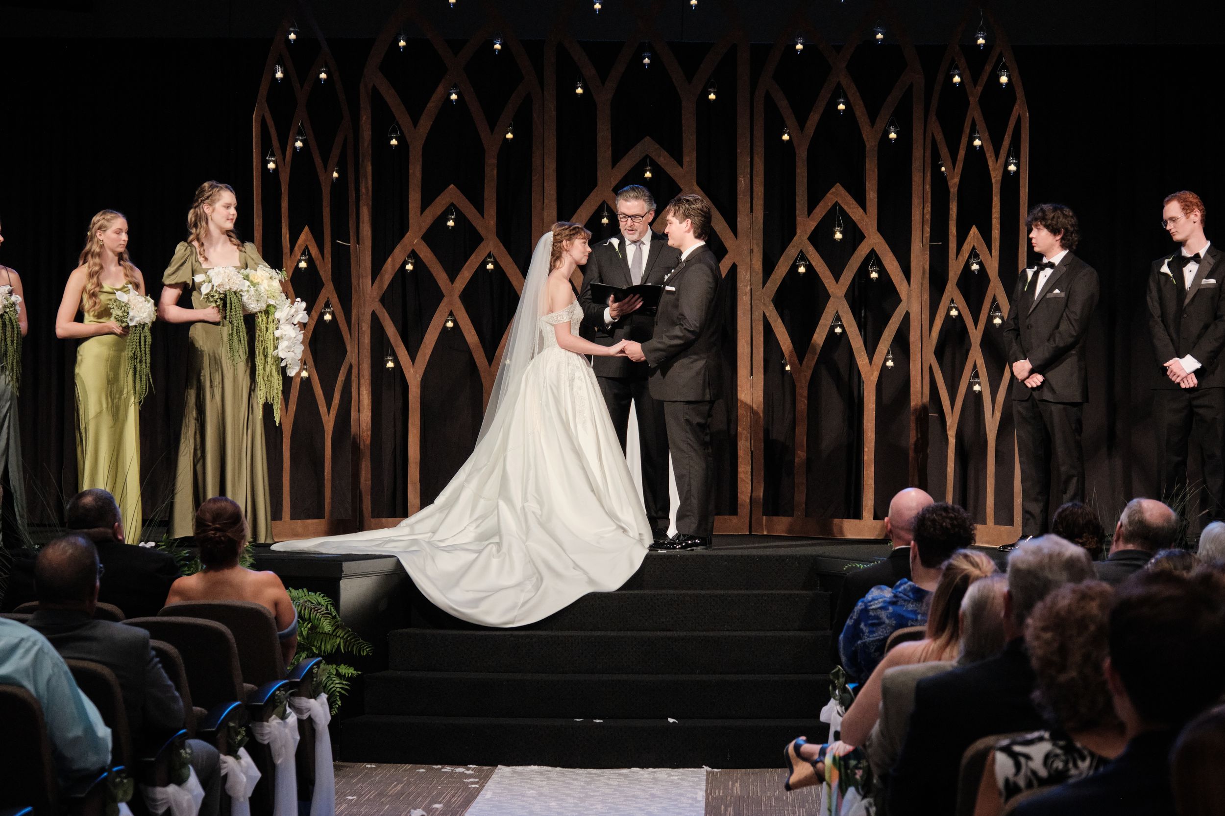 Elegant wedding ceremony with bride and groom at altar, gothic arch backdrop, bridesmaids in green gowns, guests seated in aisle.
