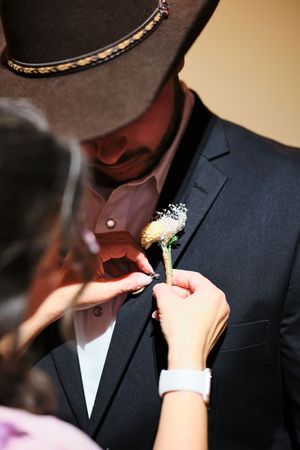 Someone pins a boutonniere flower onto a groom wearing a black suit and brown cowboy hat.