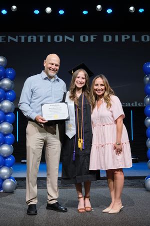 A proud graduate poses with her diploma between two smiling parents on a decorated graduation stage.