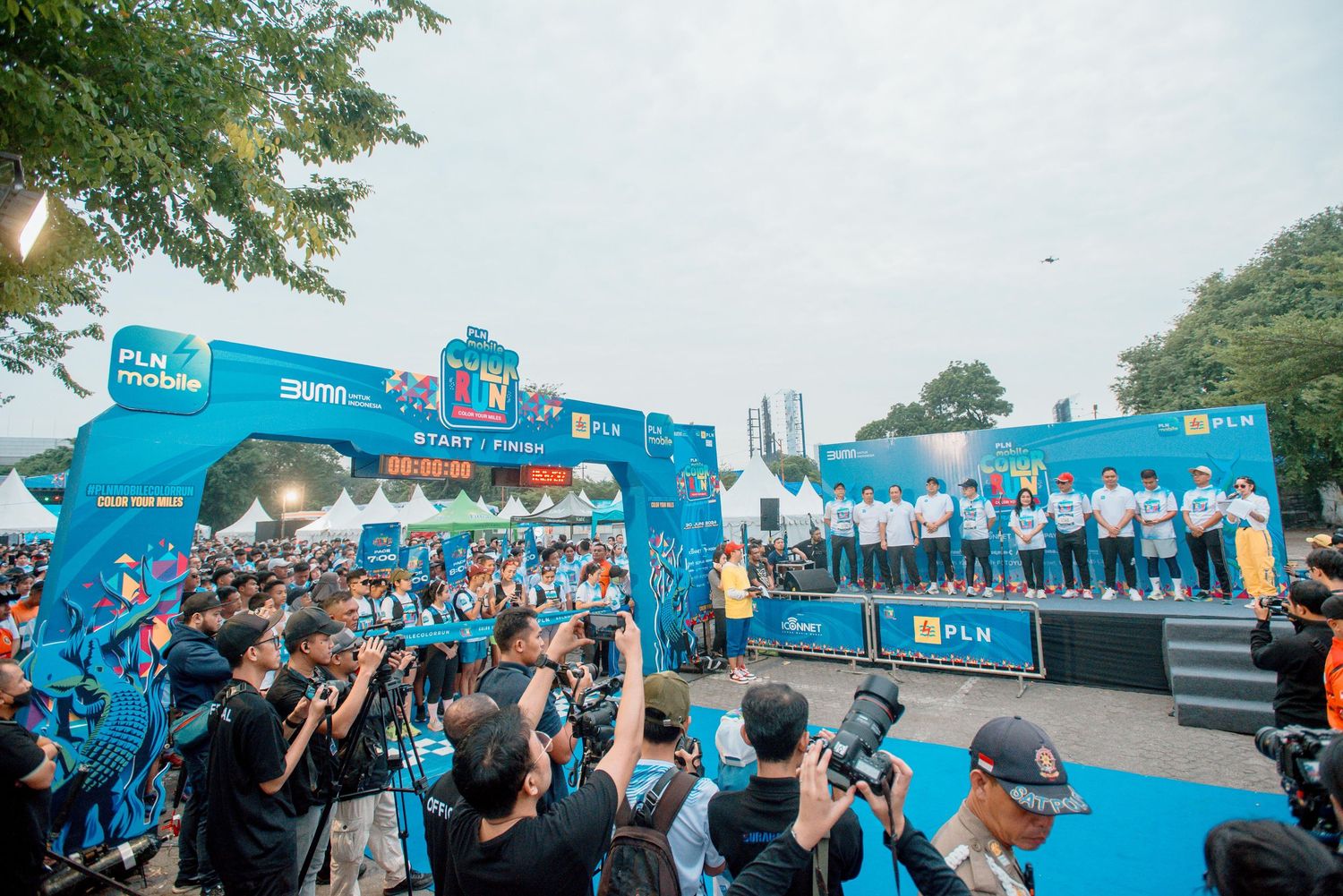 Large crowd gathers at an outdoor event with blue branded banners and signage visible in the background.
