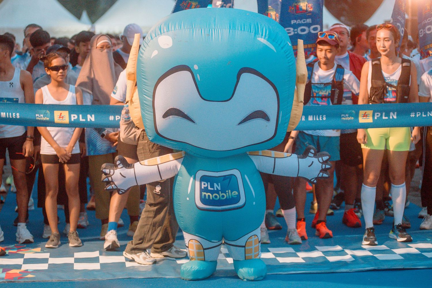 A cheerful blue mascot character with outstretched arms stands on a checkered floor at a public event.