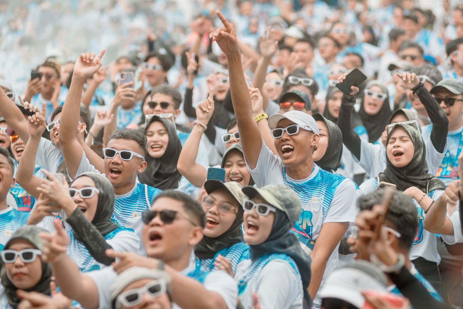 Excited sports fans in light blue jerseys and sunglasses cheer enthusiastically in packed stadium crowd.