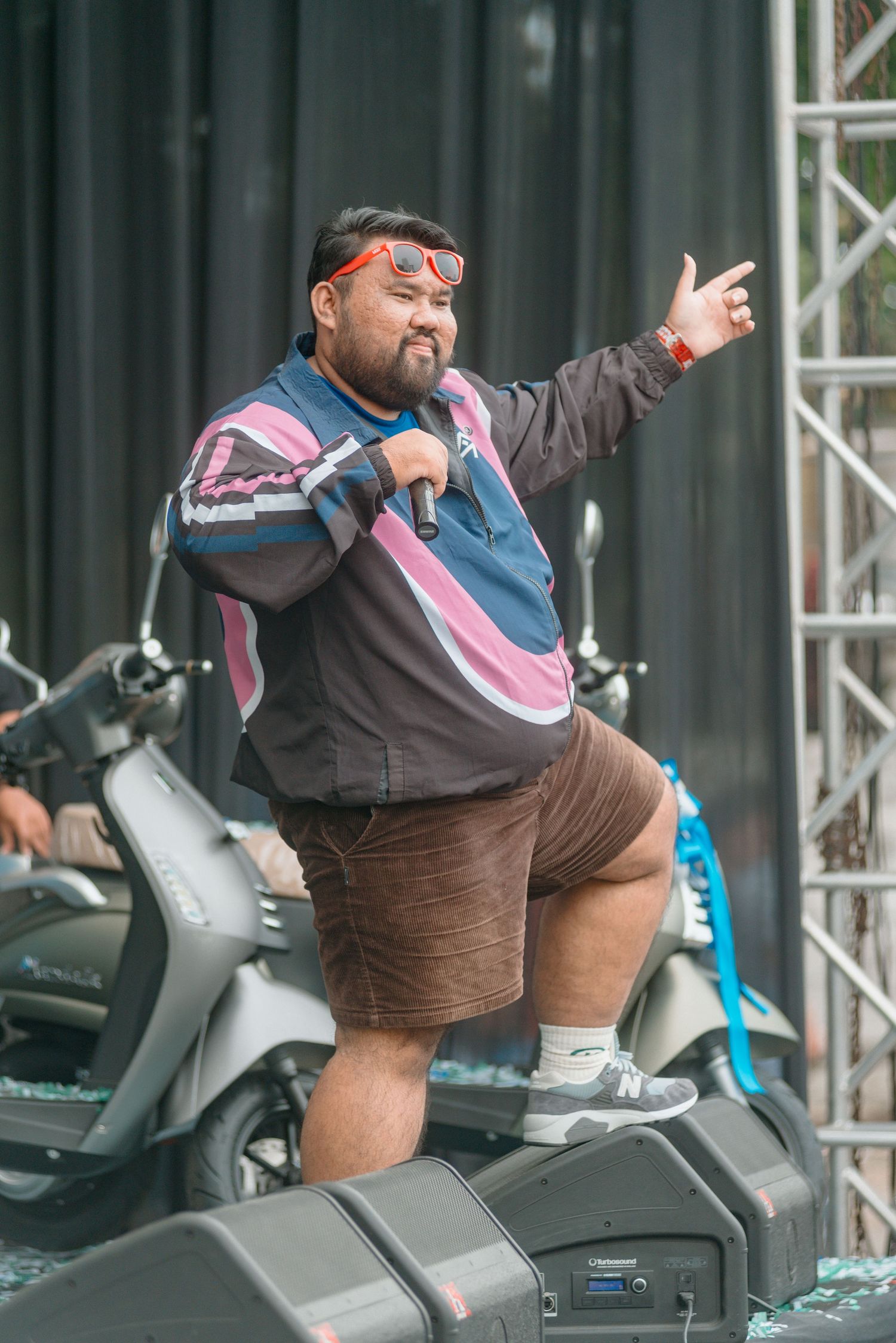 A performer in a colorful jacket and brown shorts gestures while standing on stage at an outdoor event.
