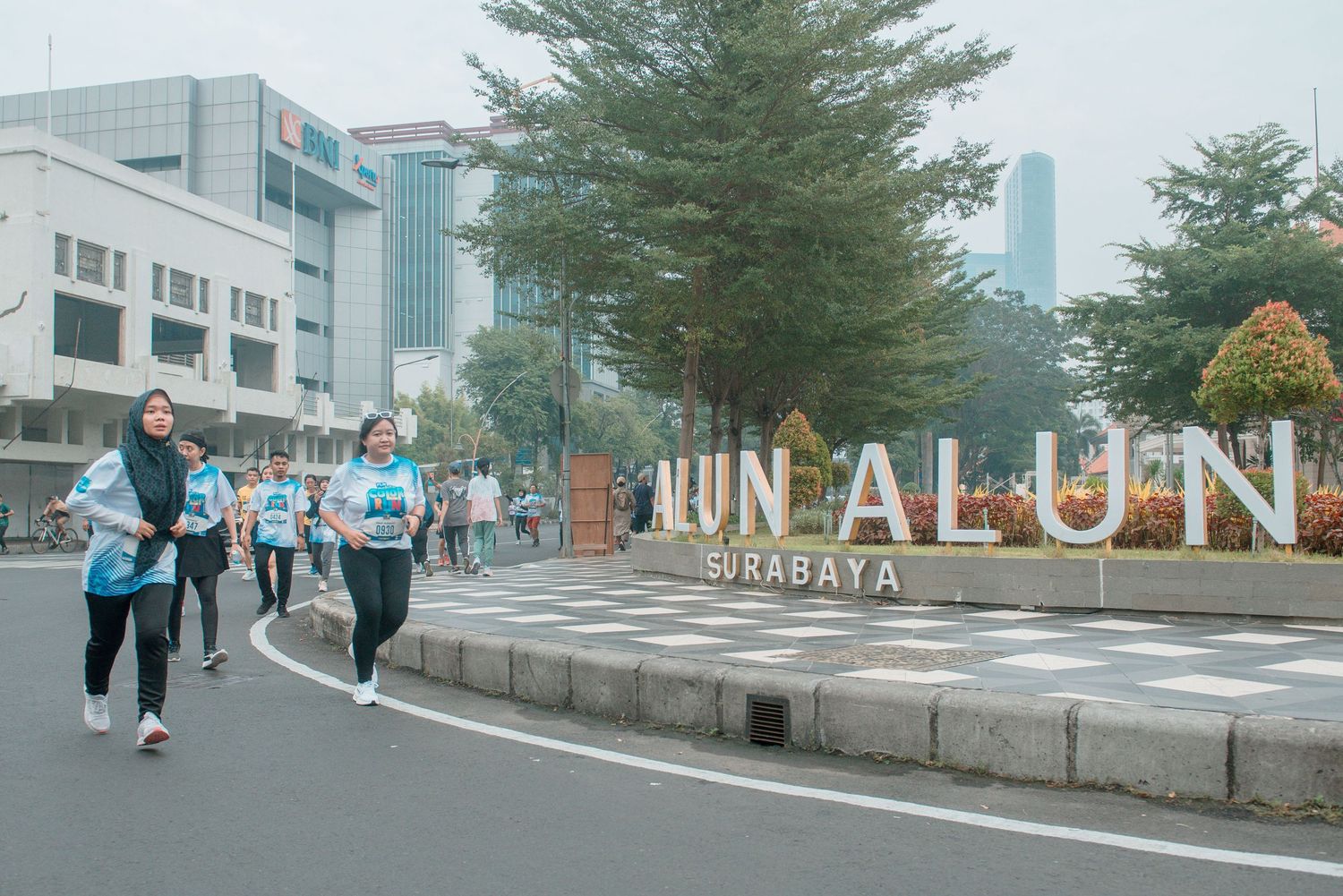 Runners jog past colorful Run Alone Surabaya sign on city street with modern buildings and trees in background.