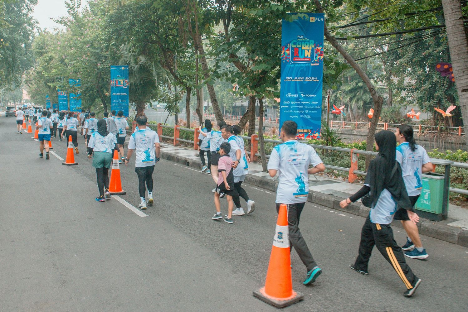 Runners in light blue shirts participate in a race along a tree-lined path with orange traffic cones marking the route.
