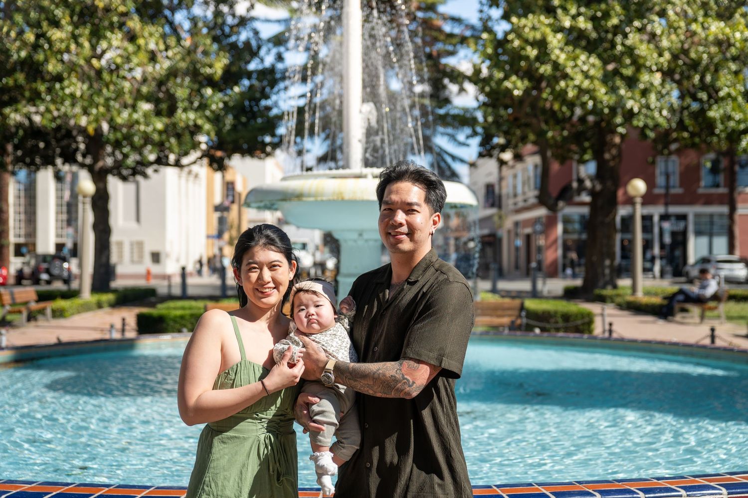 Family enjoying a sunny day together outdoors in a park setting with trees and buildings in background.