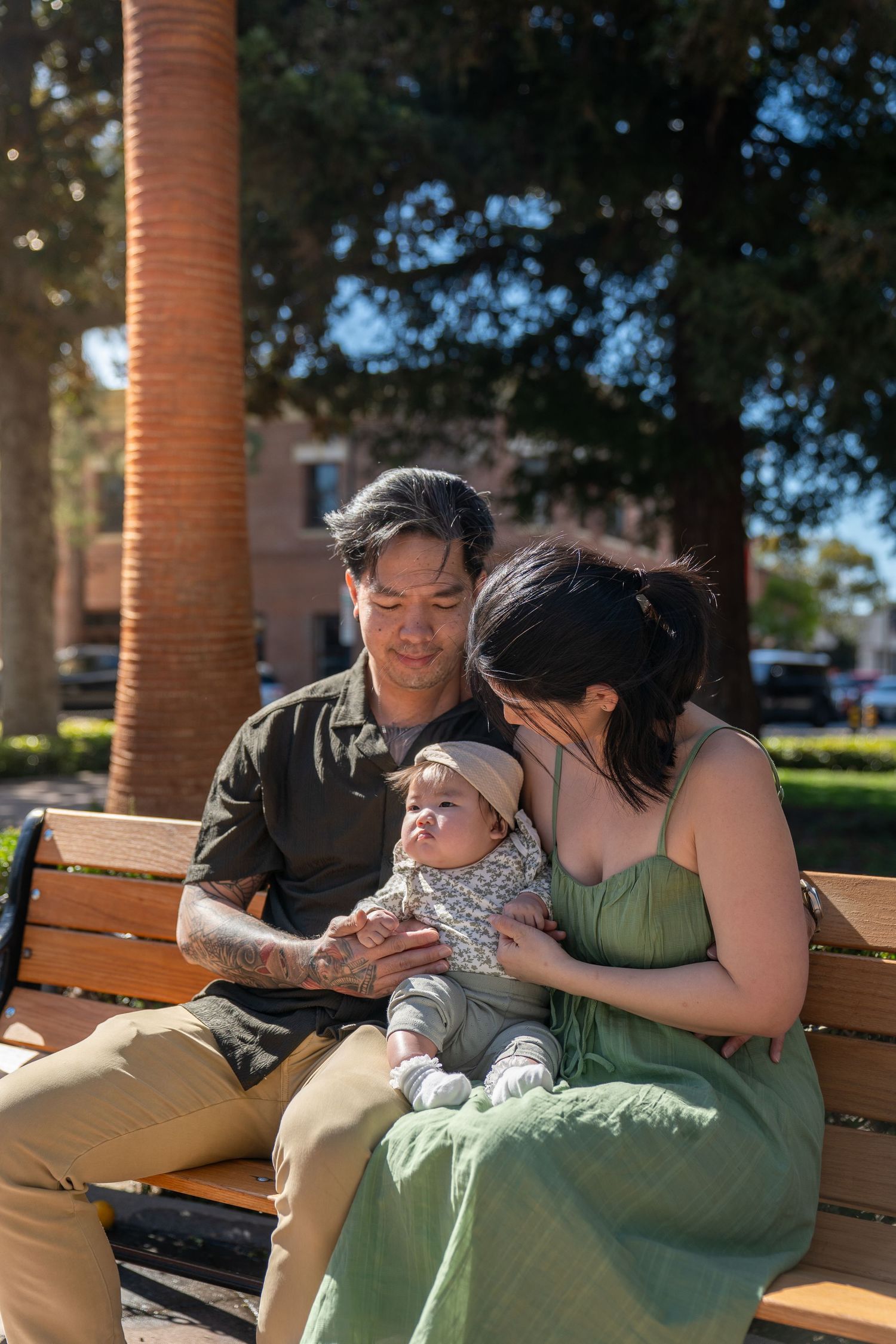 Two people share an intimate moment while sitting on a wooden bench under trees on a sunny day.