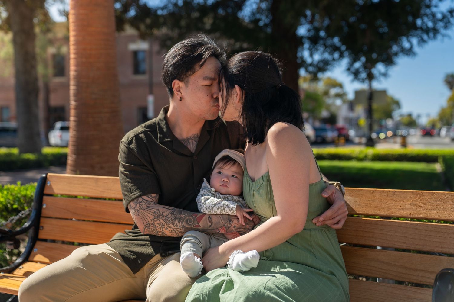 A couple embraces while sitting on a park bench with brick columns and greenery in the background.