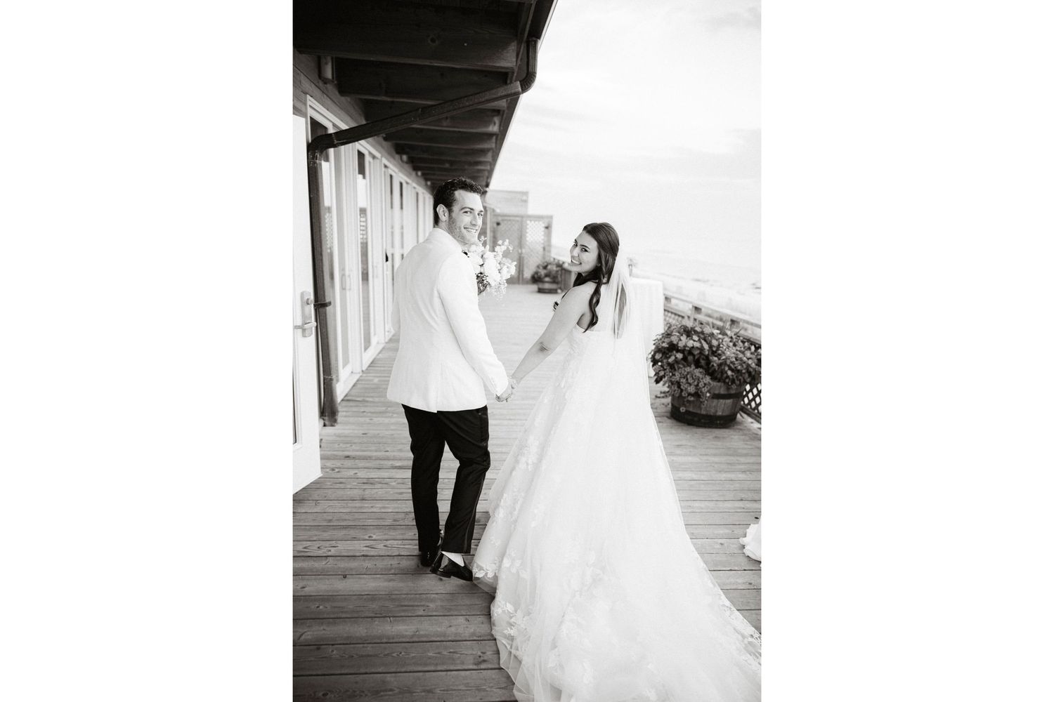 A couple in wedding attire share a romantic dance moment on a wooden deck in black and white photography.