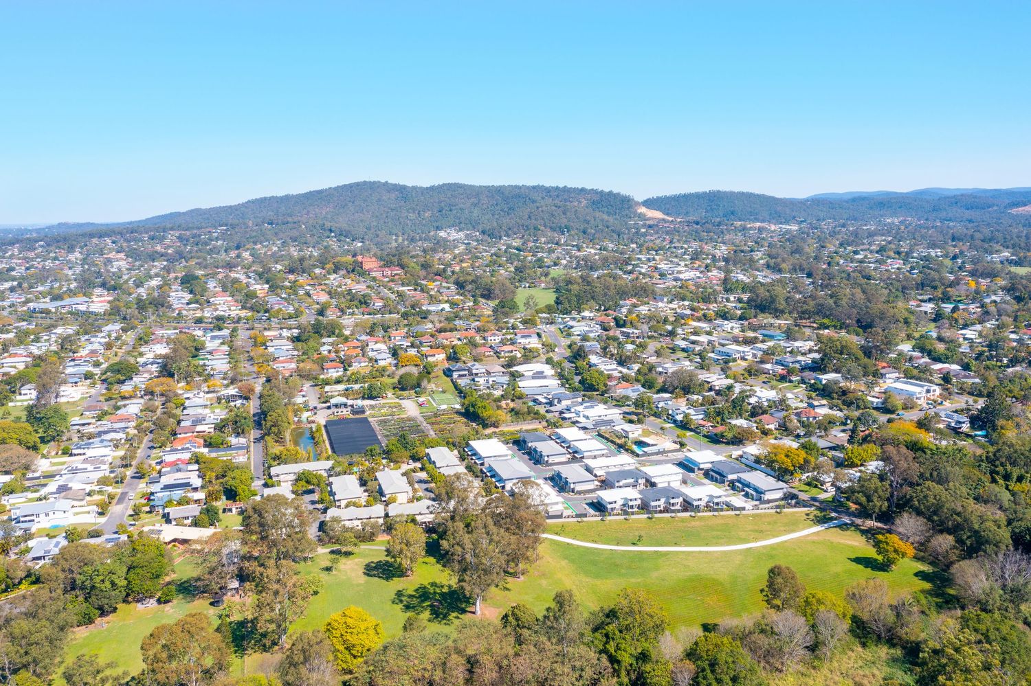 Aerial view of a leafy Brisbane suburb with a residential complex, green oval, and mountain ranges in the background.