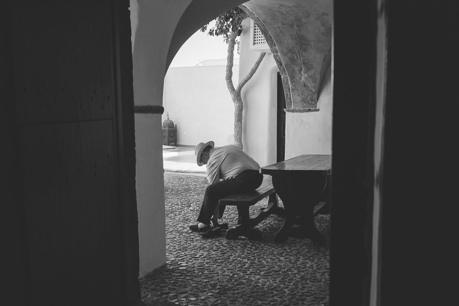 A black and white photograph shows a wedding guest sitting on a chair in a courtyard putting on his shoes viewed through an arched doorway.