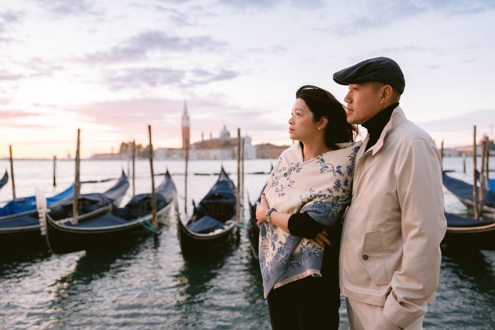 A romantic couple stands near traditional gondolas in Venice at sunset with pink and purple sky over the water.