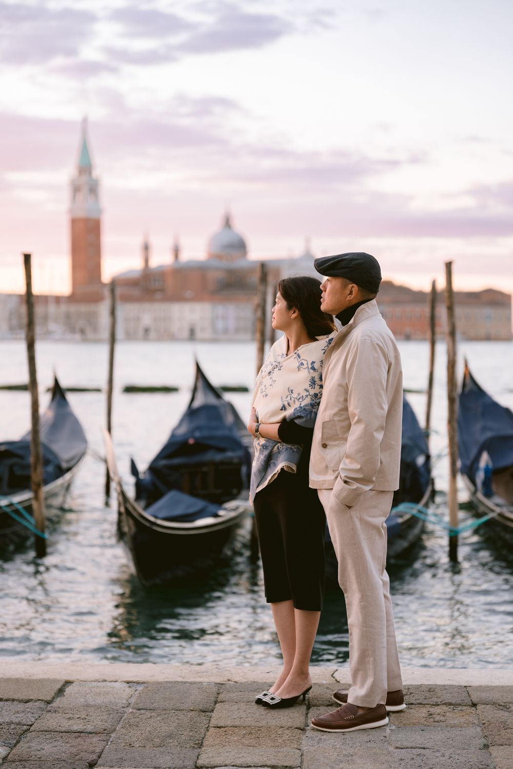 Romantic couple stands by traditional gondolas in Venice at sunset with San Giorgio Maggiore church in background.