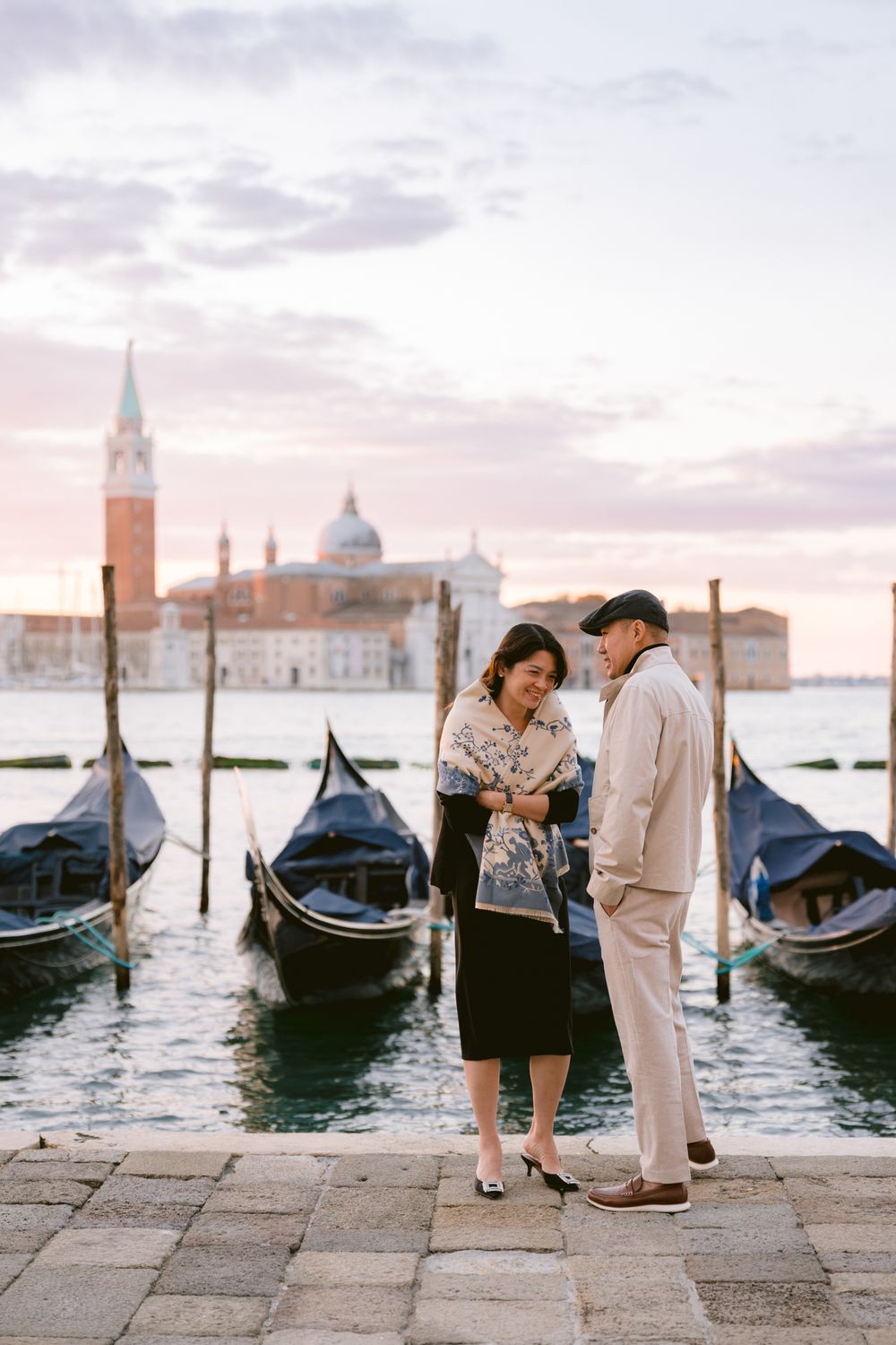 A romantic couple stands near traditional gondolas in Venice at sunset with the iconic San Giorgio Maggiore church in background.