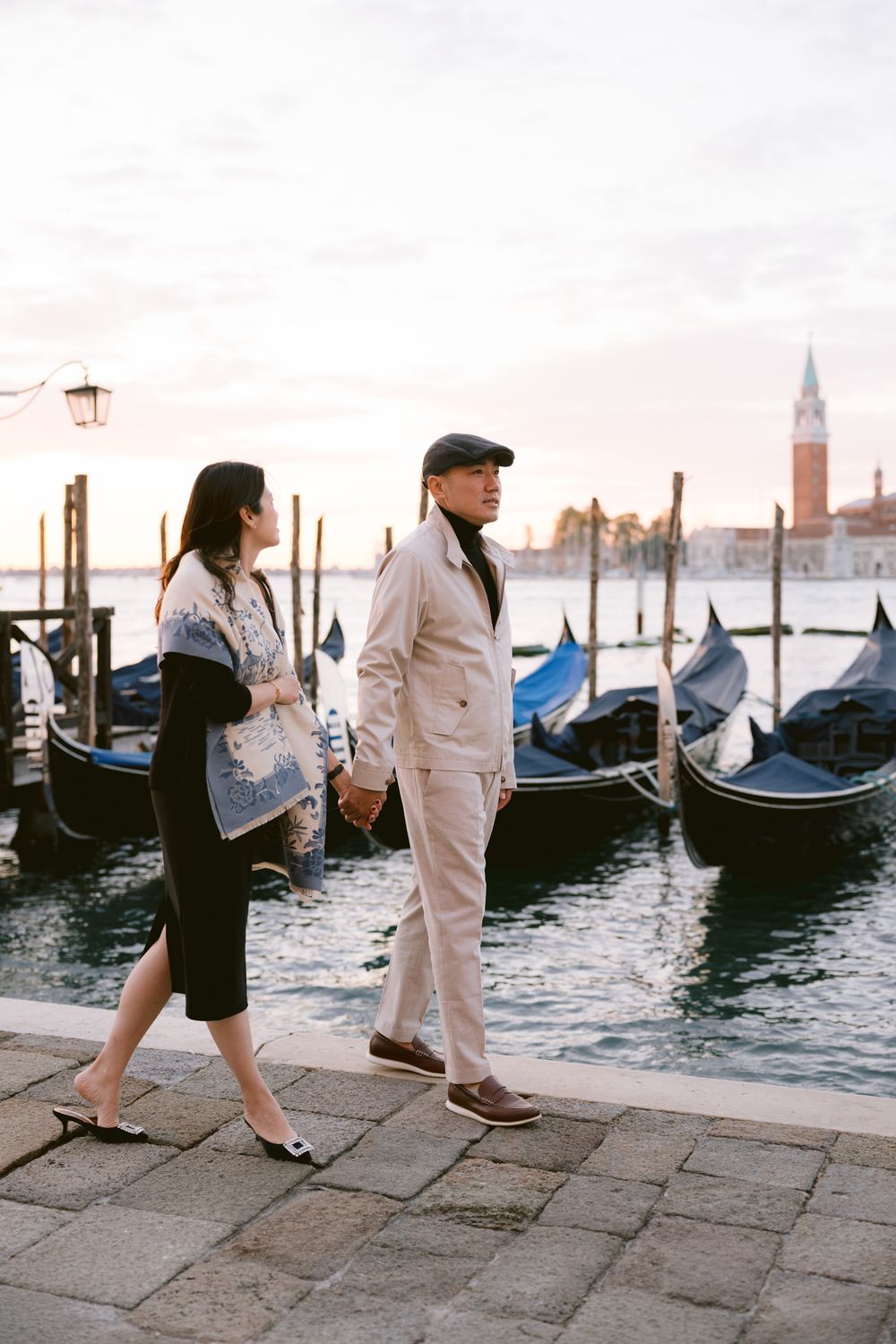 Couple walking along Venice waterfront with moored gondolas and historic skyline during golden hour.