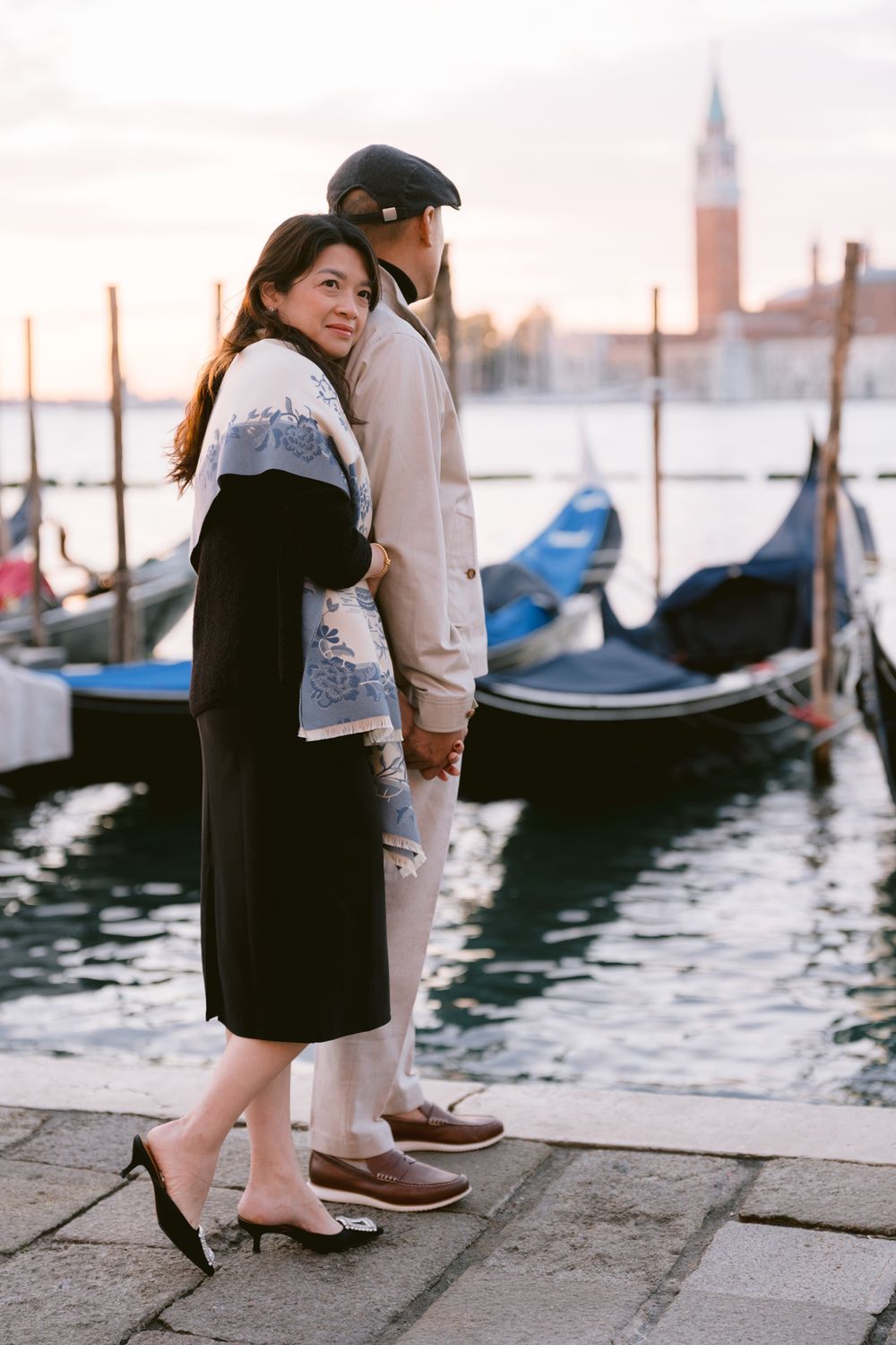 A romantic couple stands by the water's edge in Venice with traditional gondolas floating in the background at sunset.