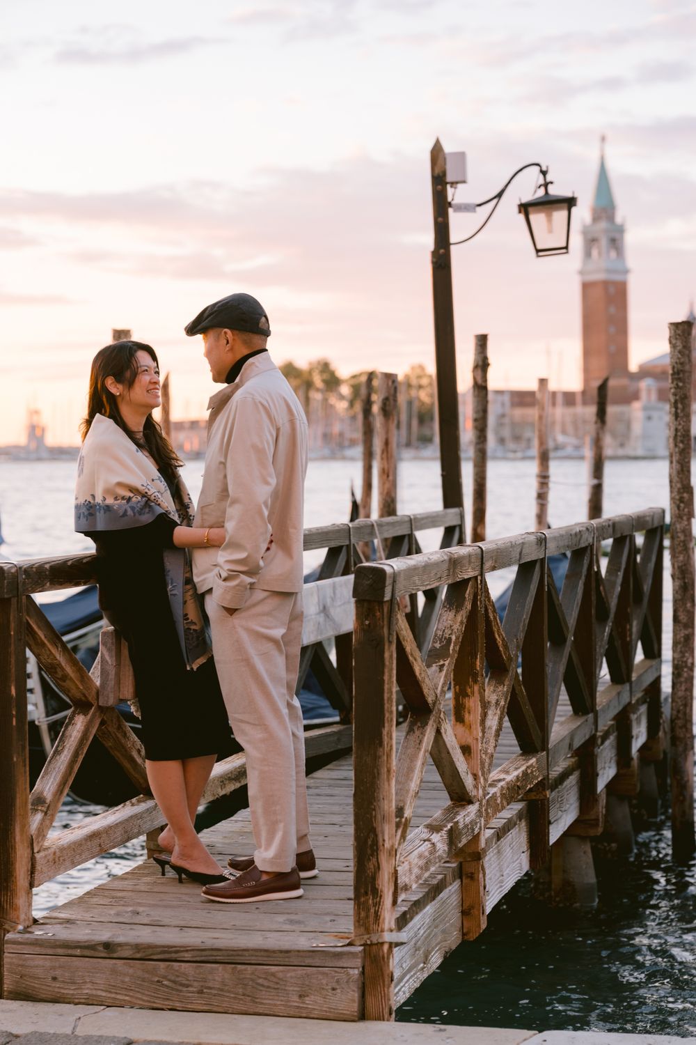 A romantic couple sits together on a wooden dock in Venice at sunset with gondolas and the city skyline in view.