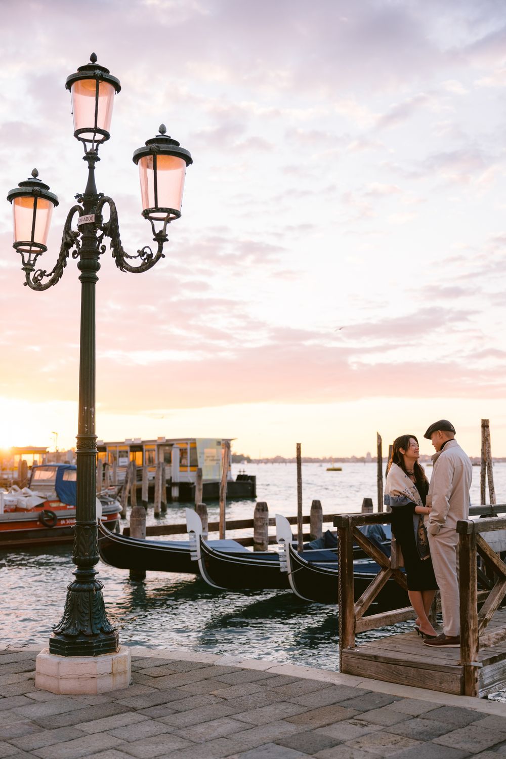 Romantic sunset view of traditional gondolas and ornate lampposts along Venice's waterfront with pink and orange sky.