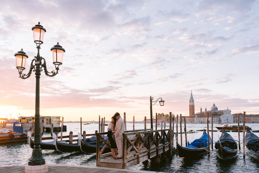 Scenic view of gondolas moored along Venice waterfront with ornate lampposts and San Giorgio Maggiore church at sunset.