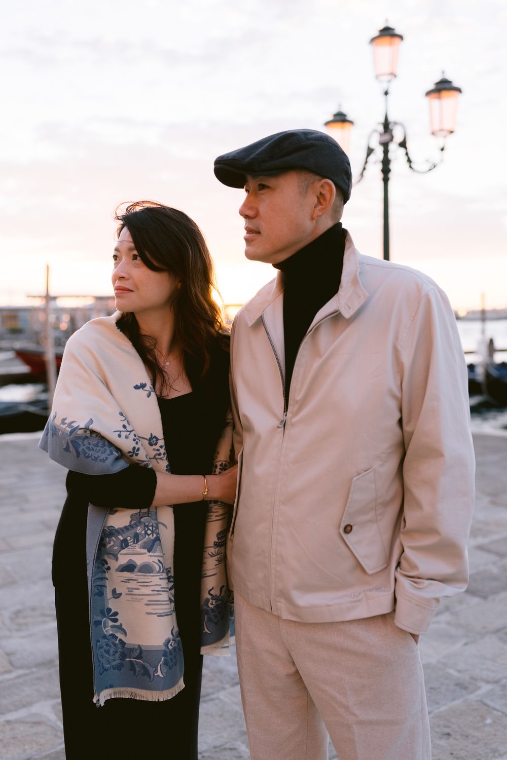 A couple poses romantically at sunset near a waterfront with vintage-style clothing and lighting.