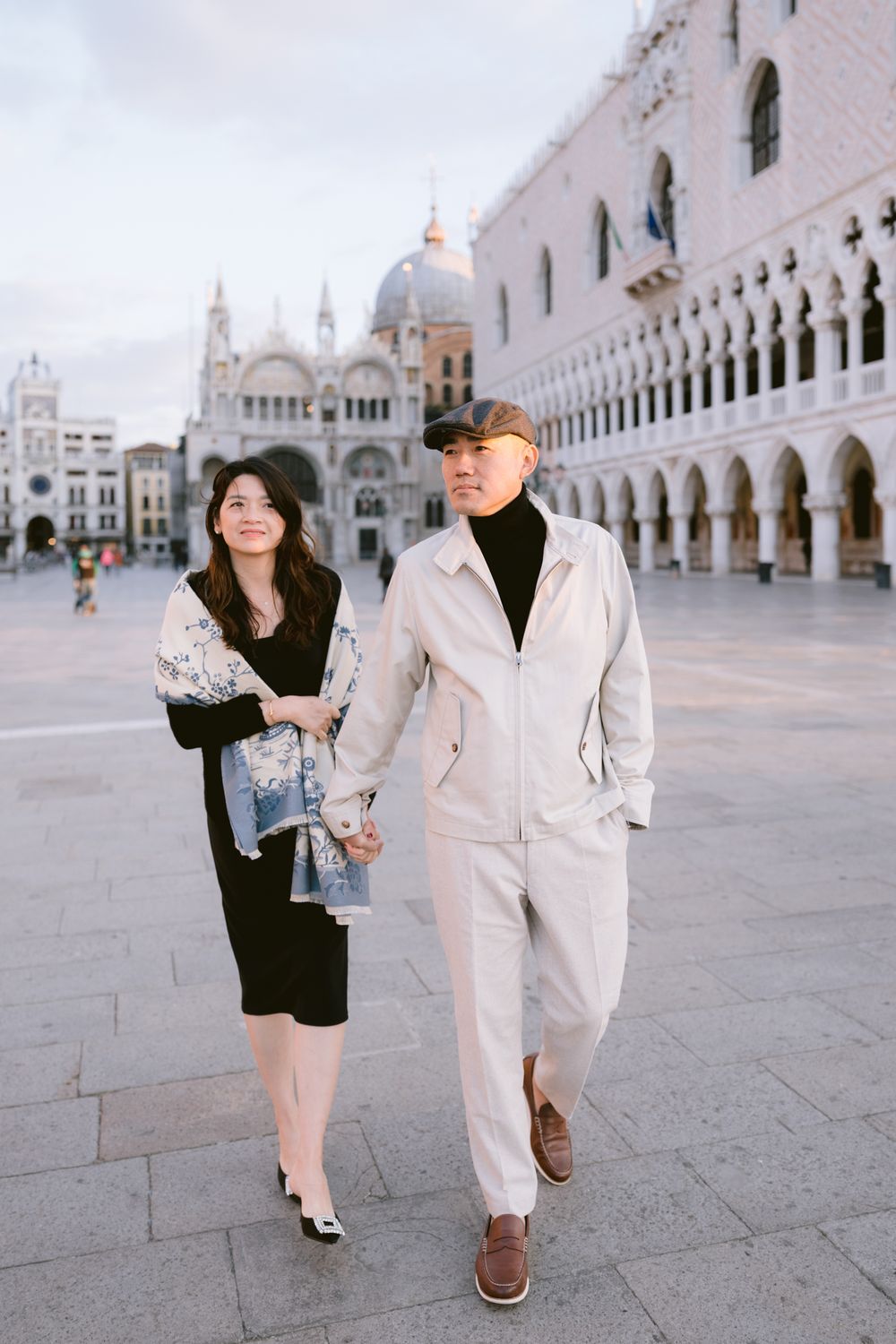 Couple walking hand in hand through Venice's iconic St. Mark's Square with classic architecture in background.
