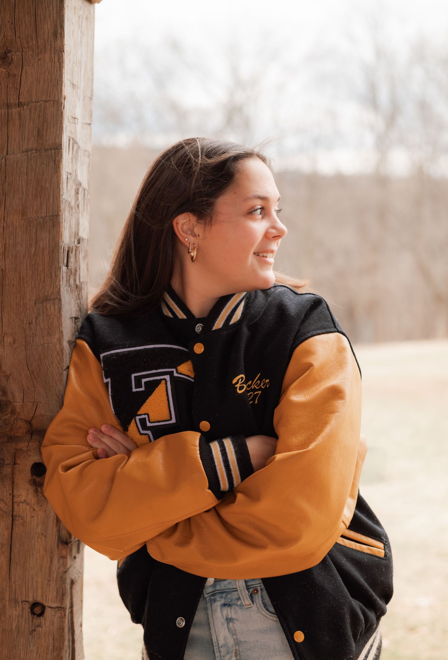 A person wearing a black and yellow varsity jacket leans against a wooden post in a natural outdoor setting.