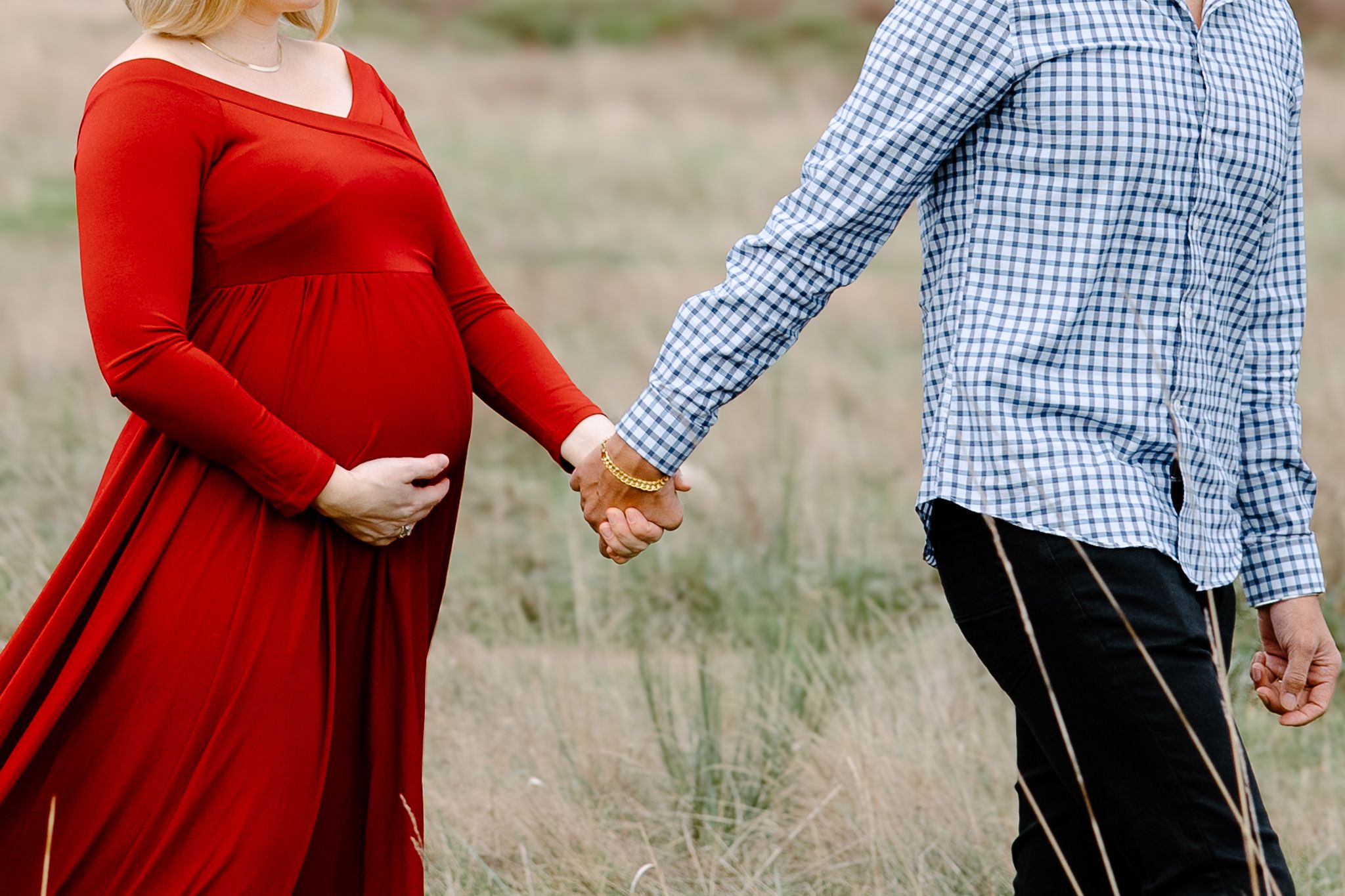 A couple in red and blue gingham outfits hold hands while walking through a grassy meadow.
