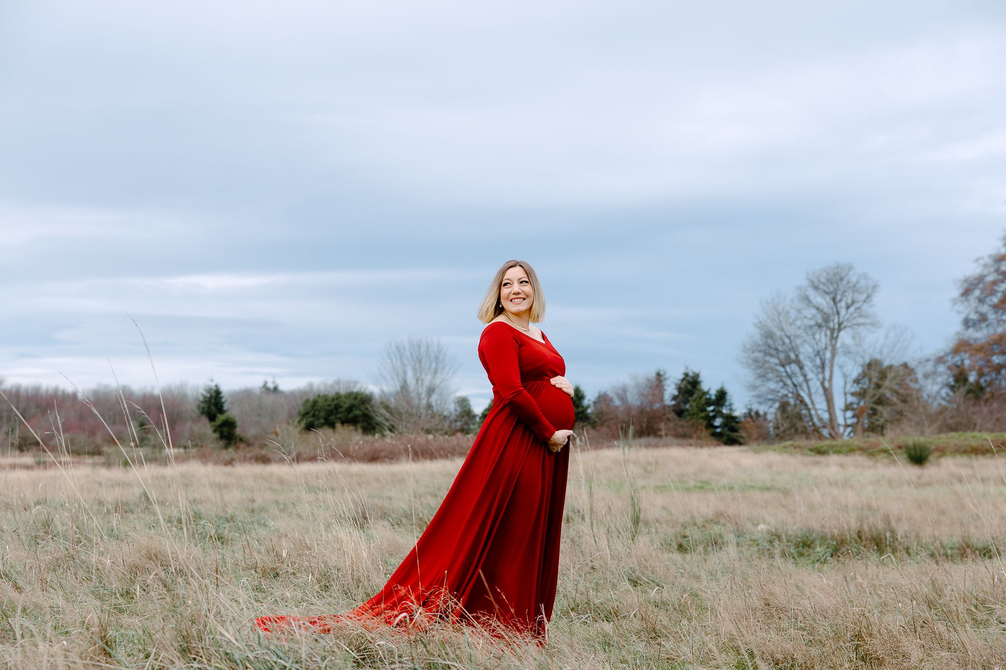 A figure in a flowing red dress stands in a serene autumn field under cloudy skies.