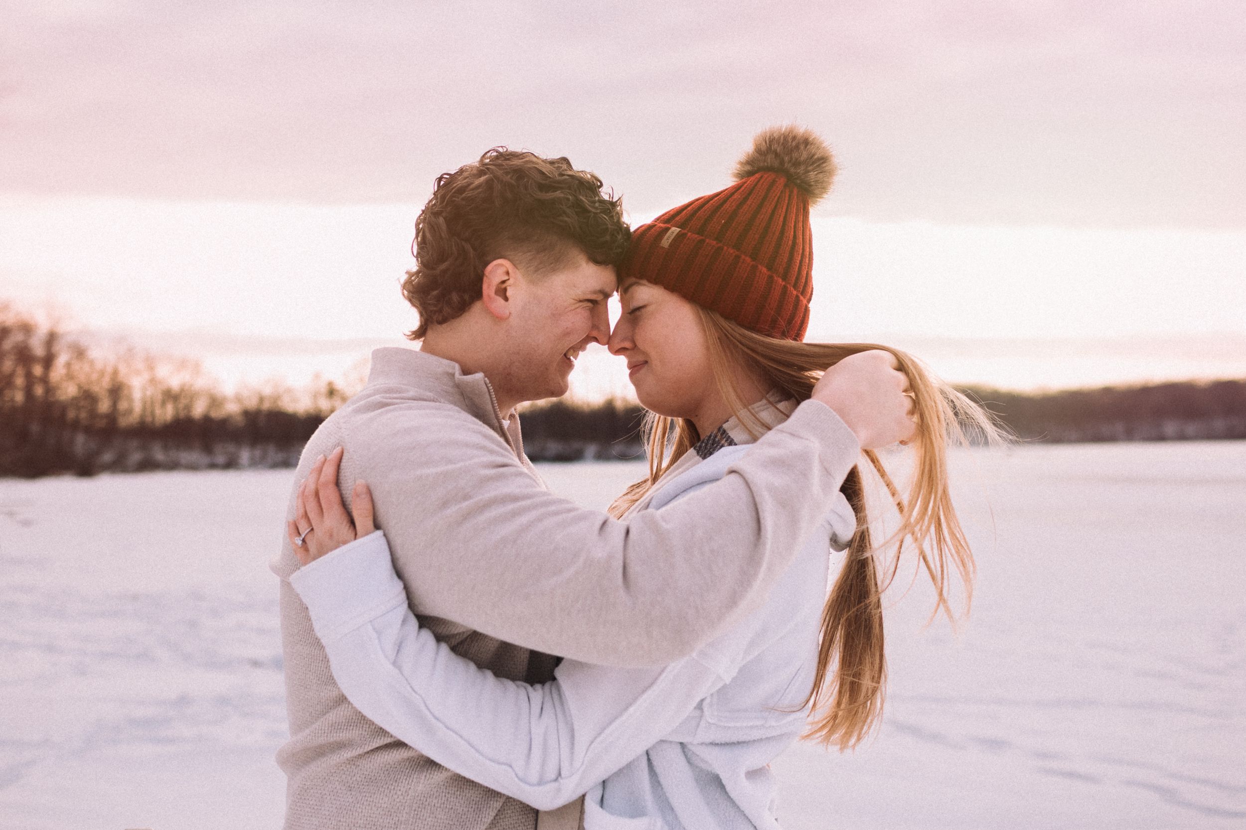 A couple embraces in a romantic moment against a snowy winter landscape at sunset.