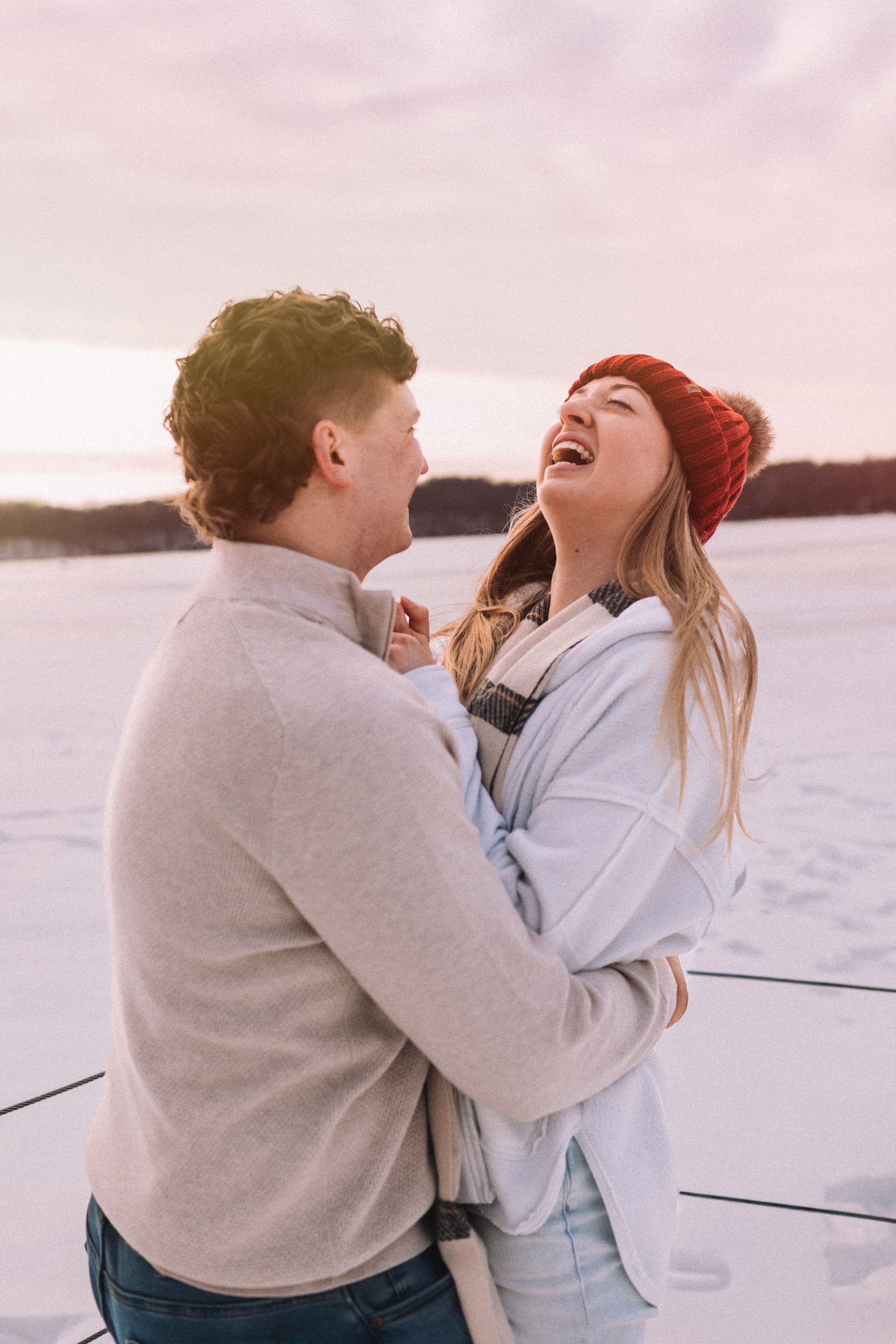 A couple shares a joyful embrace and laugh together while standing near a snowy lake on a winter day.