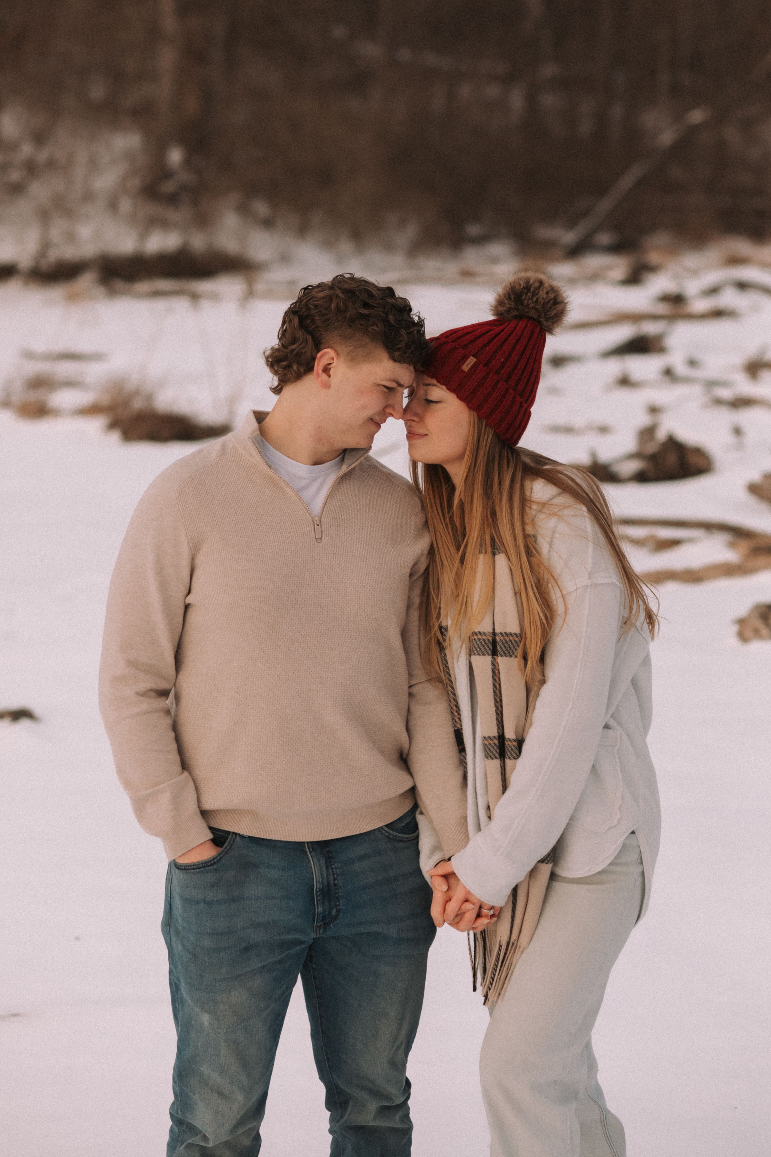 A couple shares an intimate moment in the snow wearing cozy winter clothing including a red beanie and neutral sweaters.