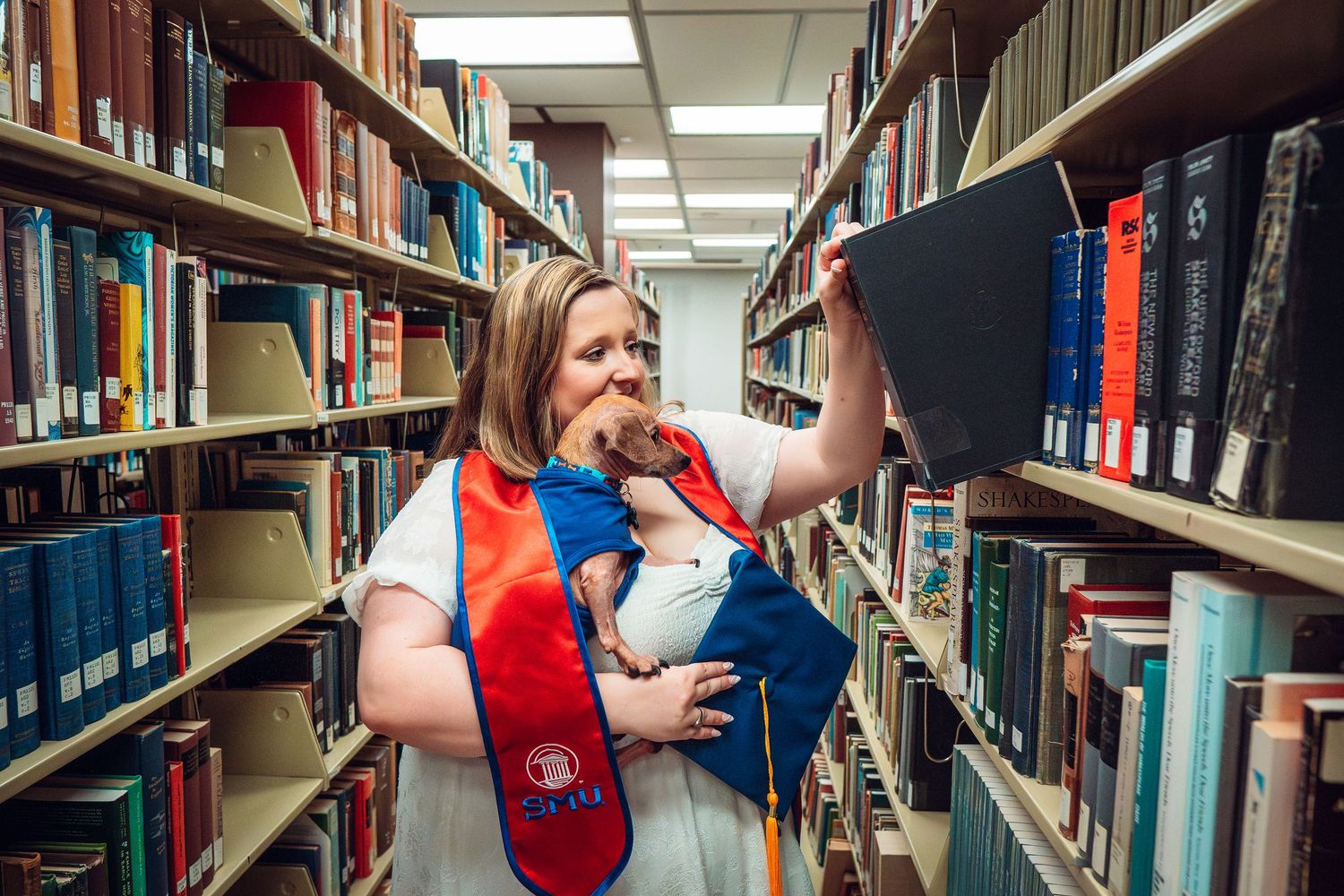 Graduate in red stole playfully poses with books in library stacks.