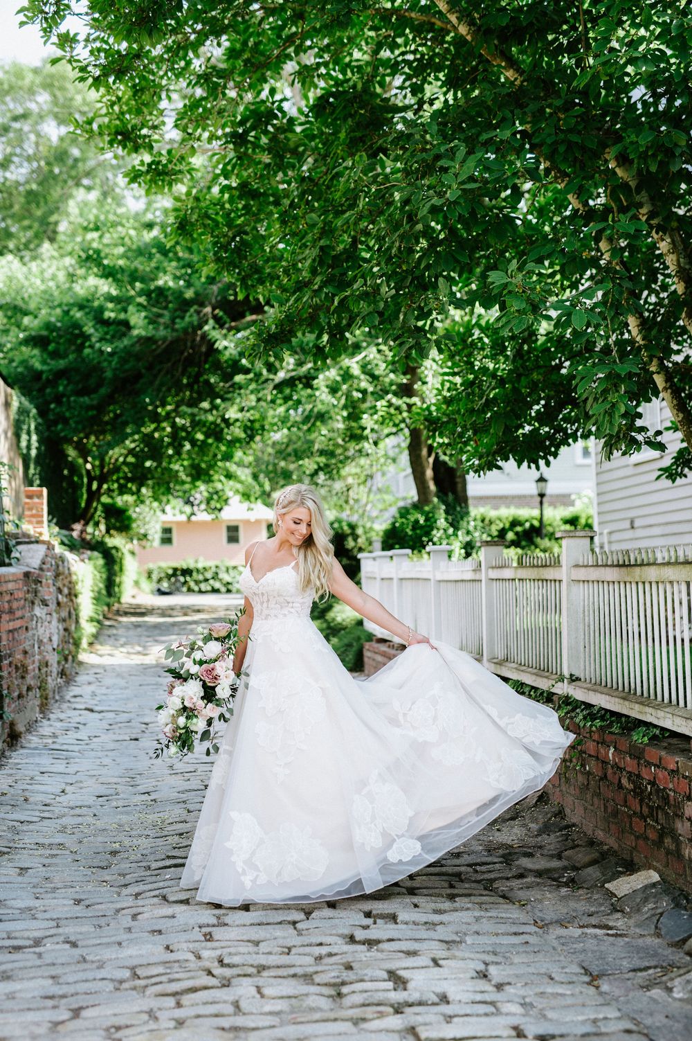 A bride in a flowing white wedding dress twirls joyfully on a cobblestone path surrounded by lush green trees.