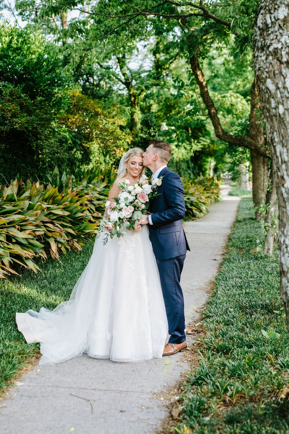 A romantic wedding couple shares an intimate moment on a tree-lined garden path with lush greenery.