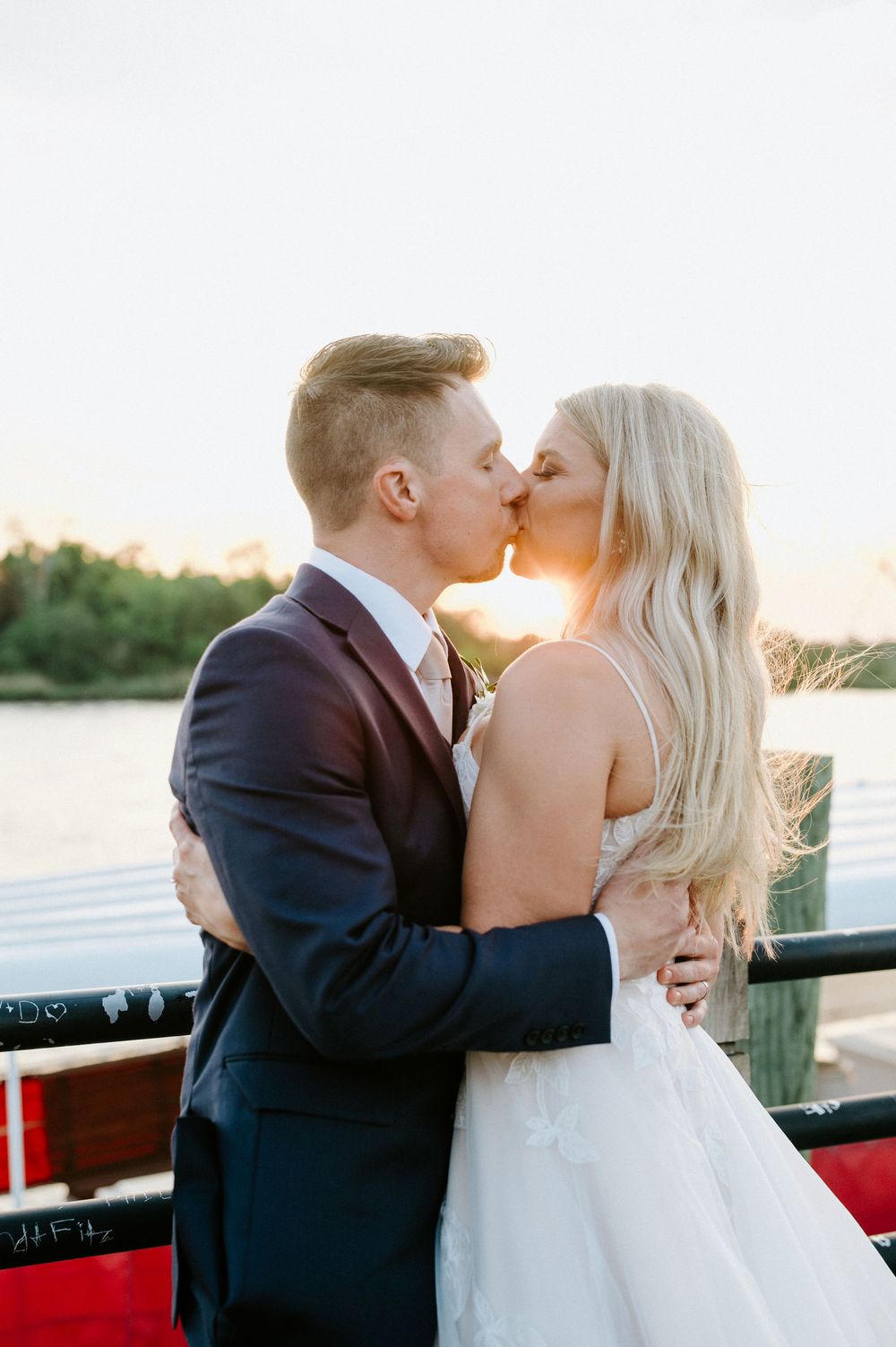 Romantic sunset wedding photos of a couple kissing on a waterfront deck in formal attire.