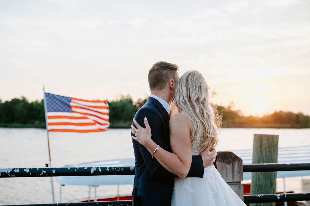 A couple shares a romantic moment at sunset with an American flag waving in the background near a dock.