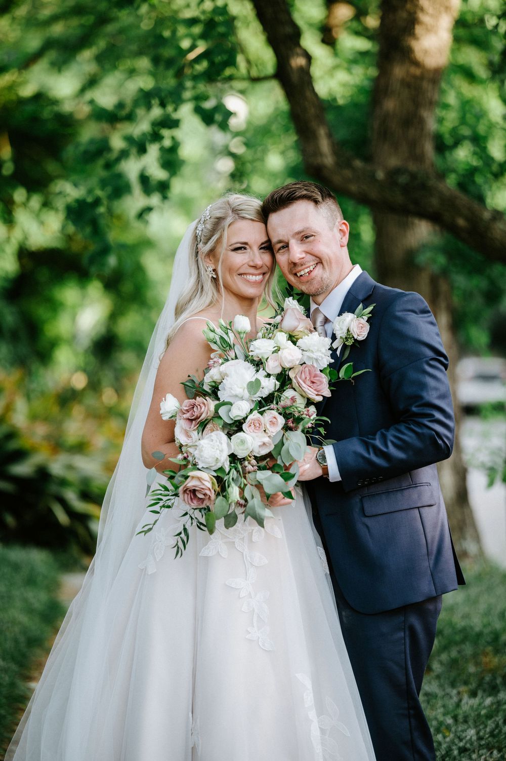 Newlyweds pose for romantic portraits in a lush garden setting with white and pink floral bouquet.