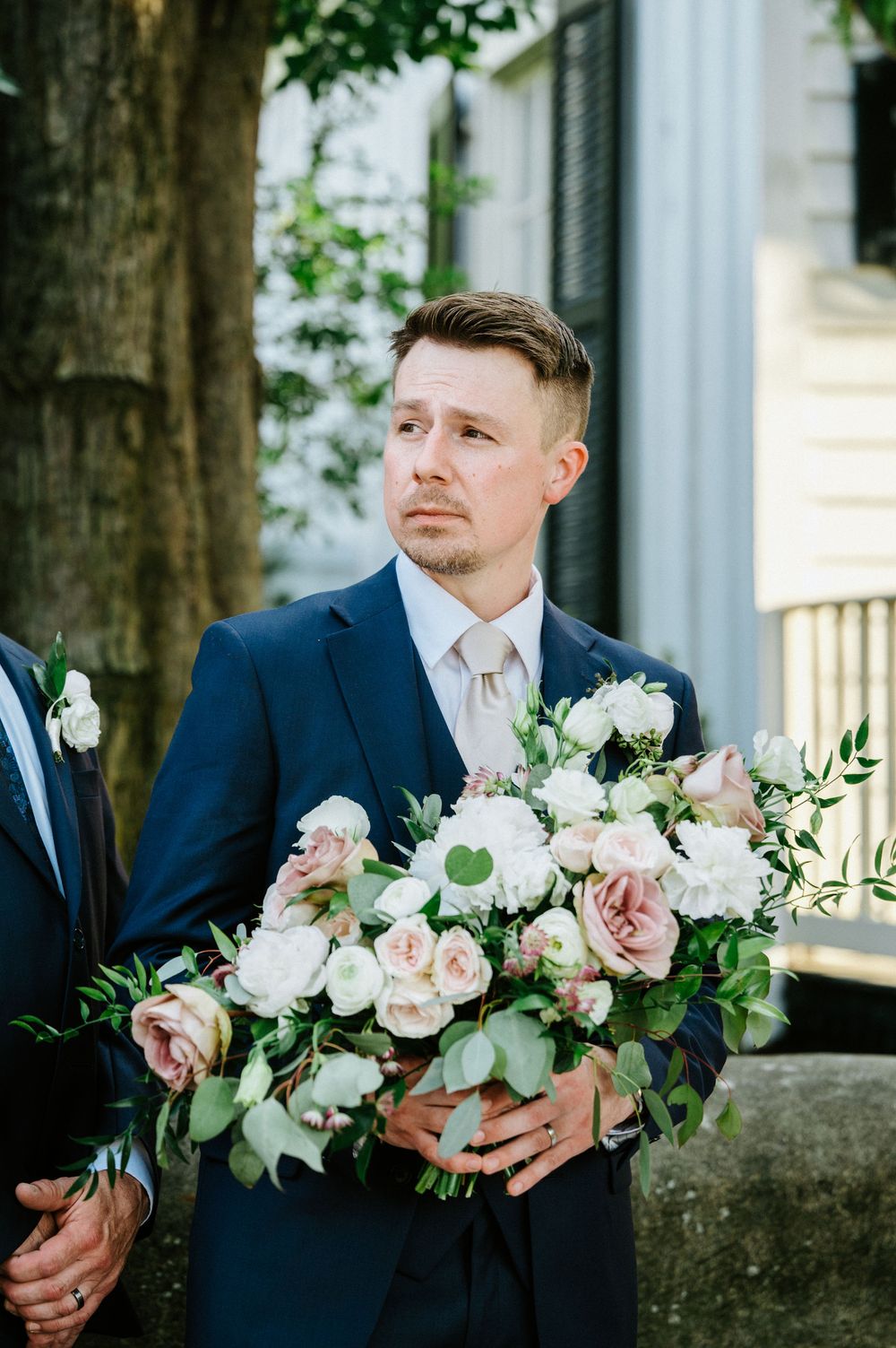 Groom in navy suit holds elegant bouquet of white and blush roses with greenery.