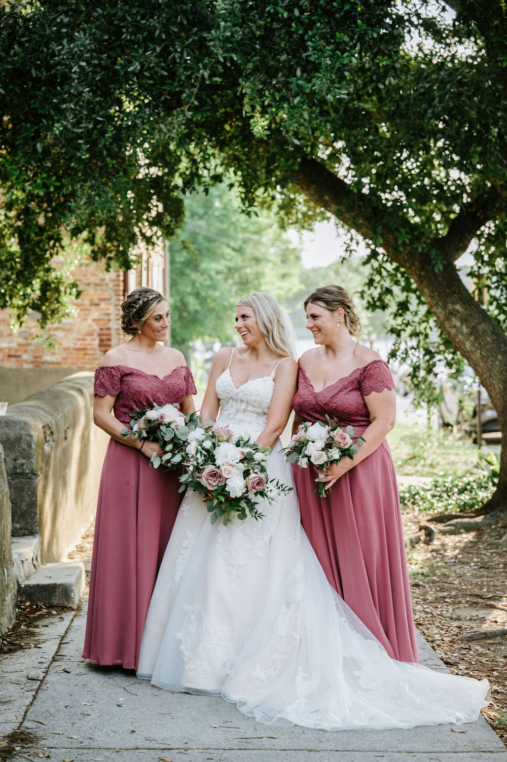 Bride in white gown poses with bridesmaids wearing dusty rose dresses under shaded trees.