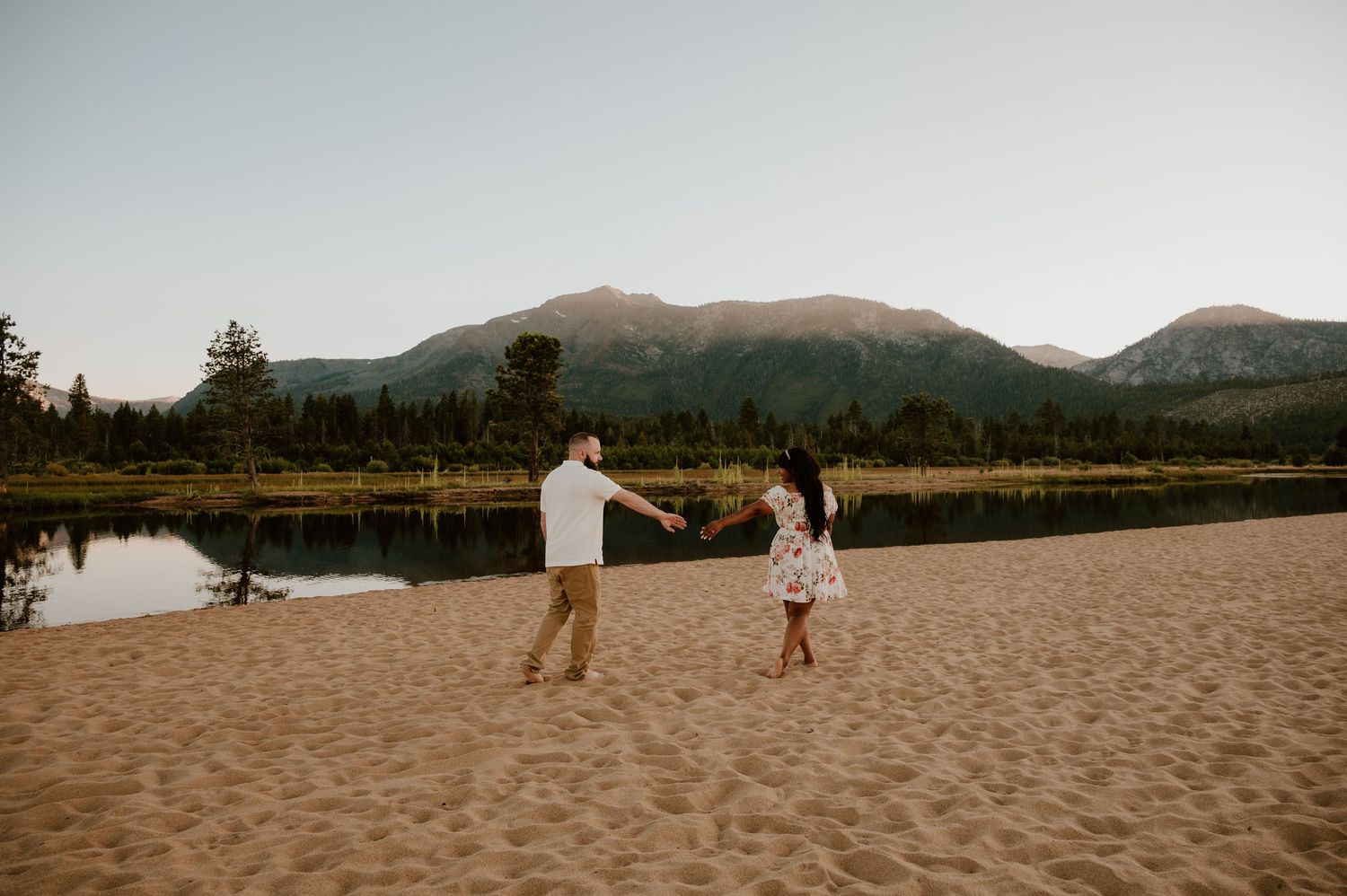 A couple walks along a scenic sandy beach at sunset with mountains and water in the background.