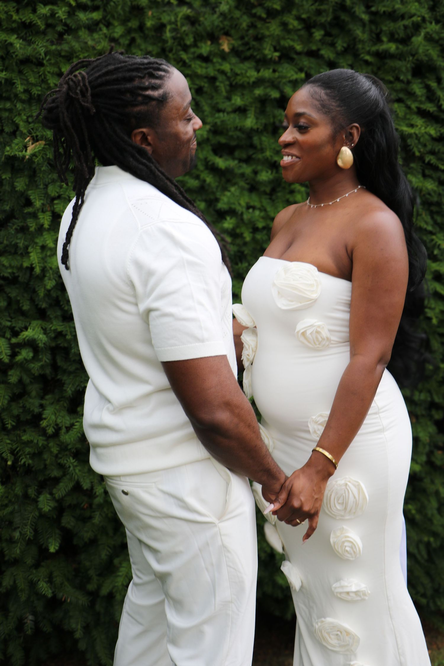 A couple in matching white outfits hold hands and gaze at each other against a green hedge backdrop.