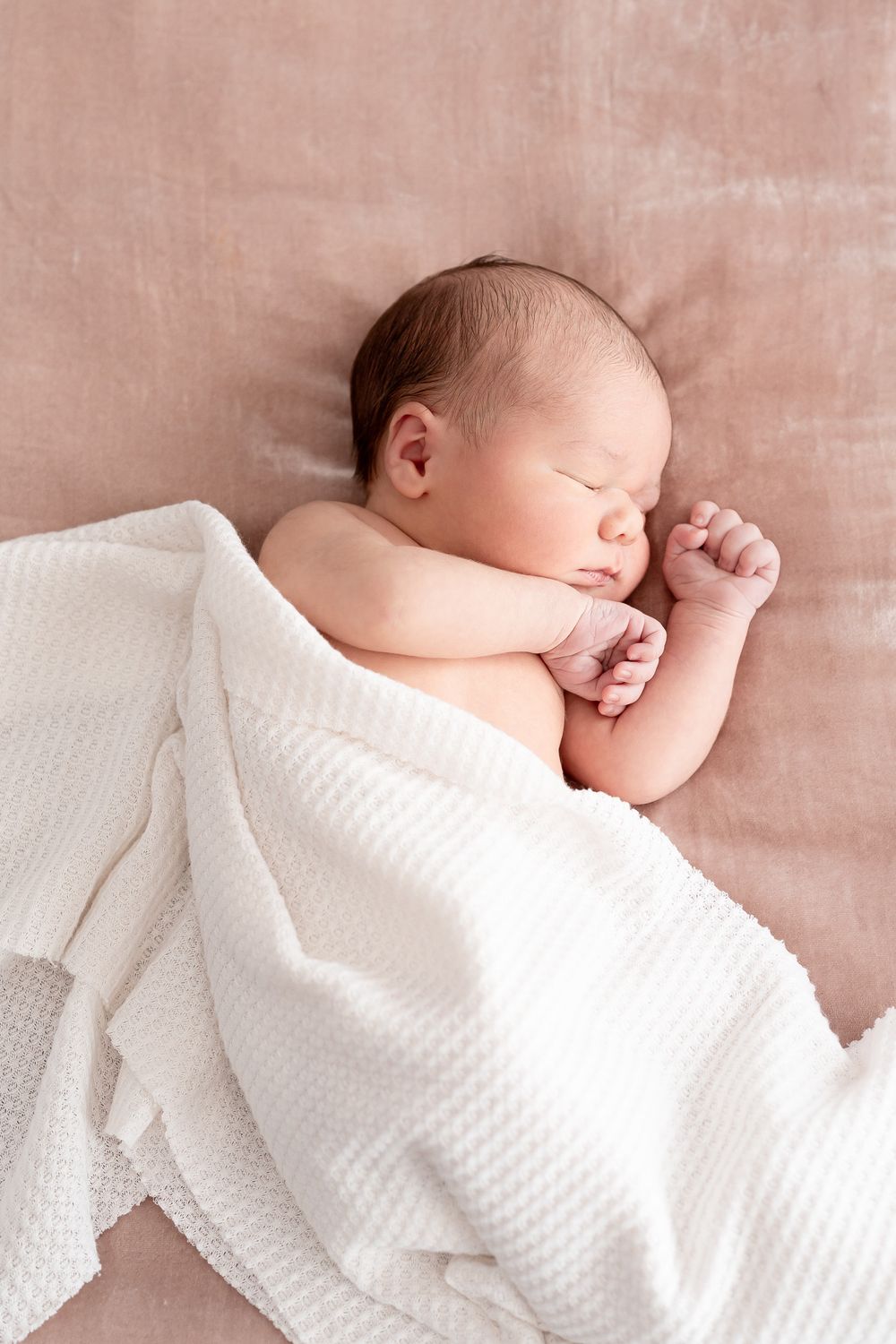 Peaceful sleeping newborn baby wrapped partially in soft white textured blanket against pink background.