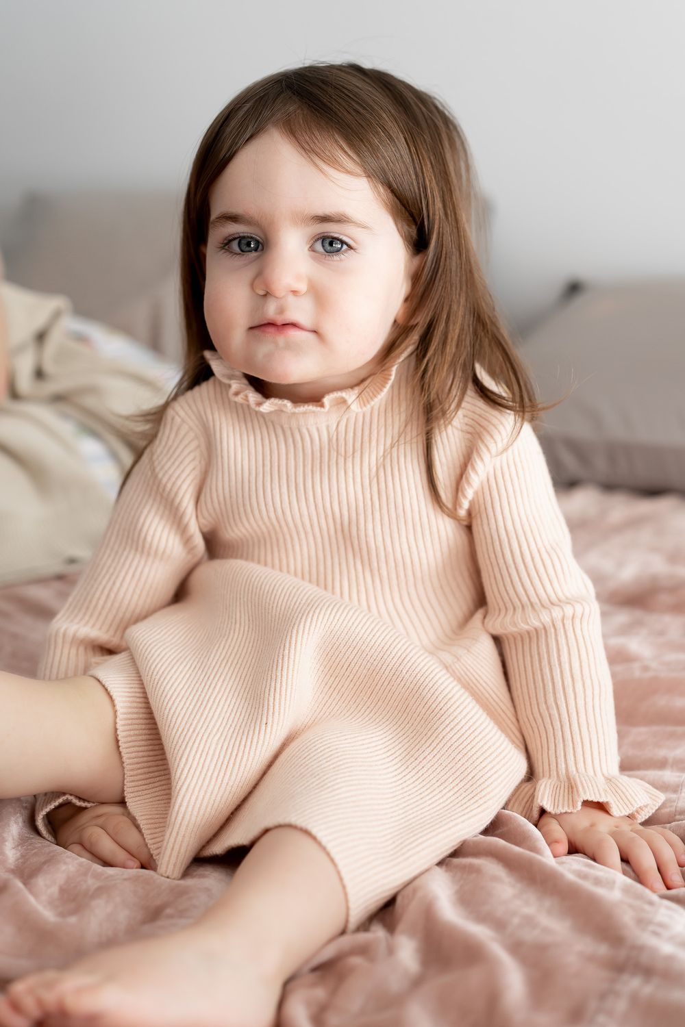 Young child in beige ribbed sweater dress sitting on pink bedding.