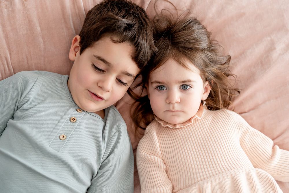 Two children in soft pastel outfits cuddle together while resting on a blush pink background.
