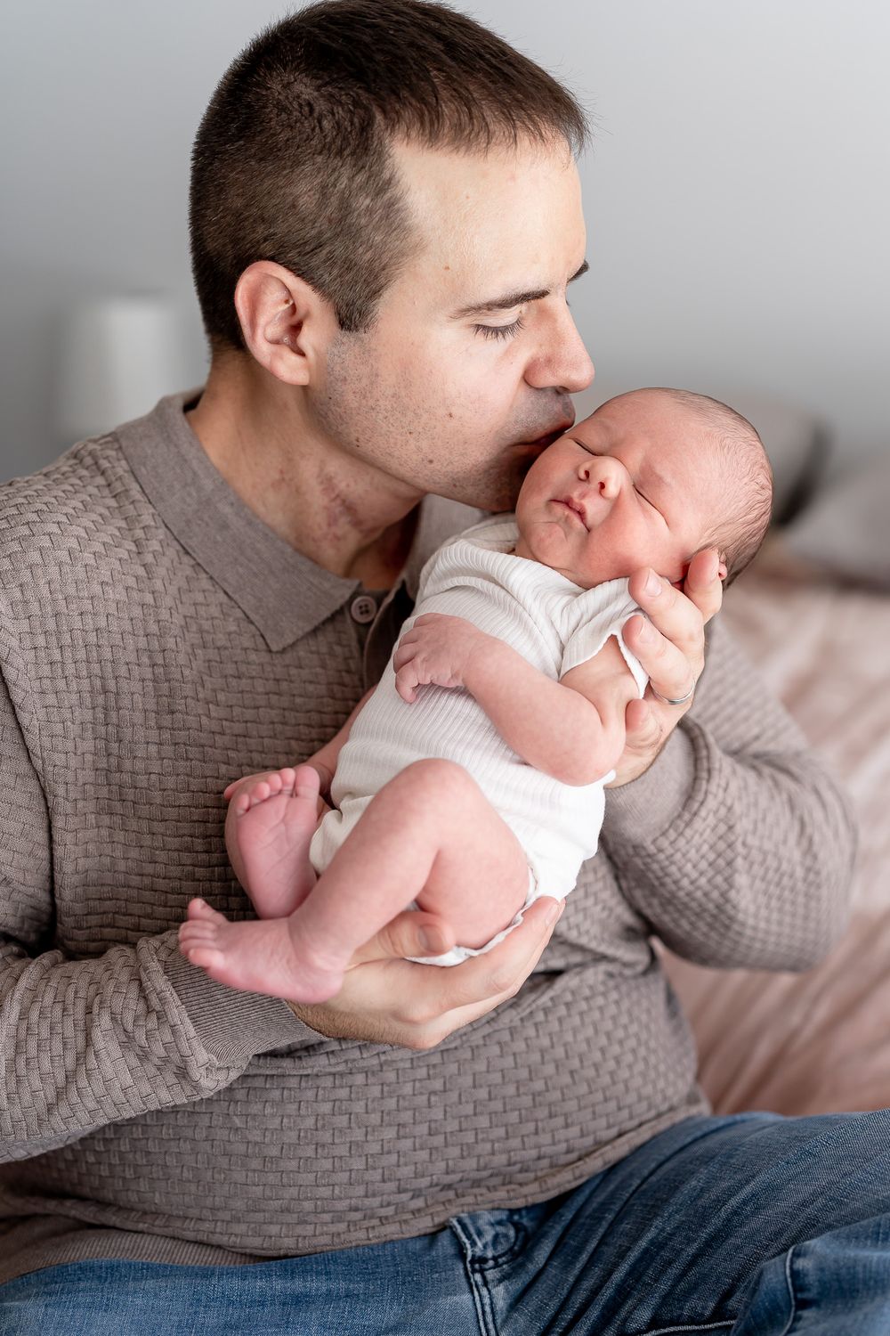 A tender moment between parent and newborn baby in a grey sweater holding their infant close and giving a gentle kiss.