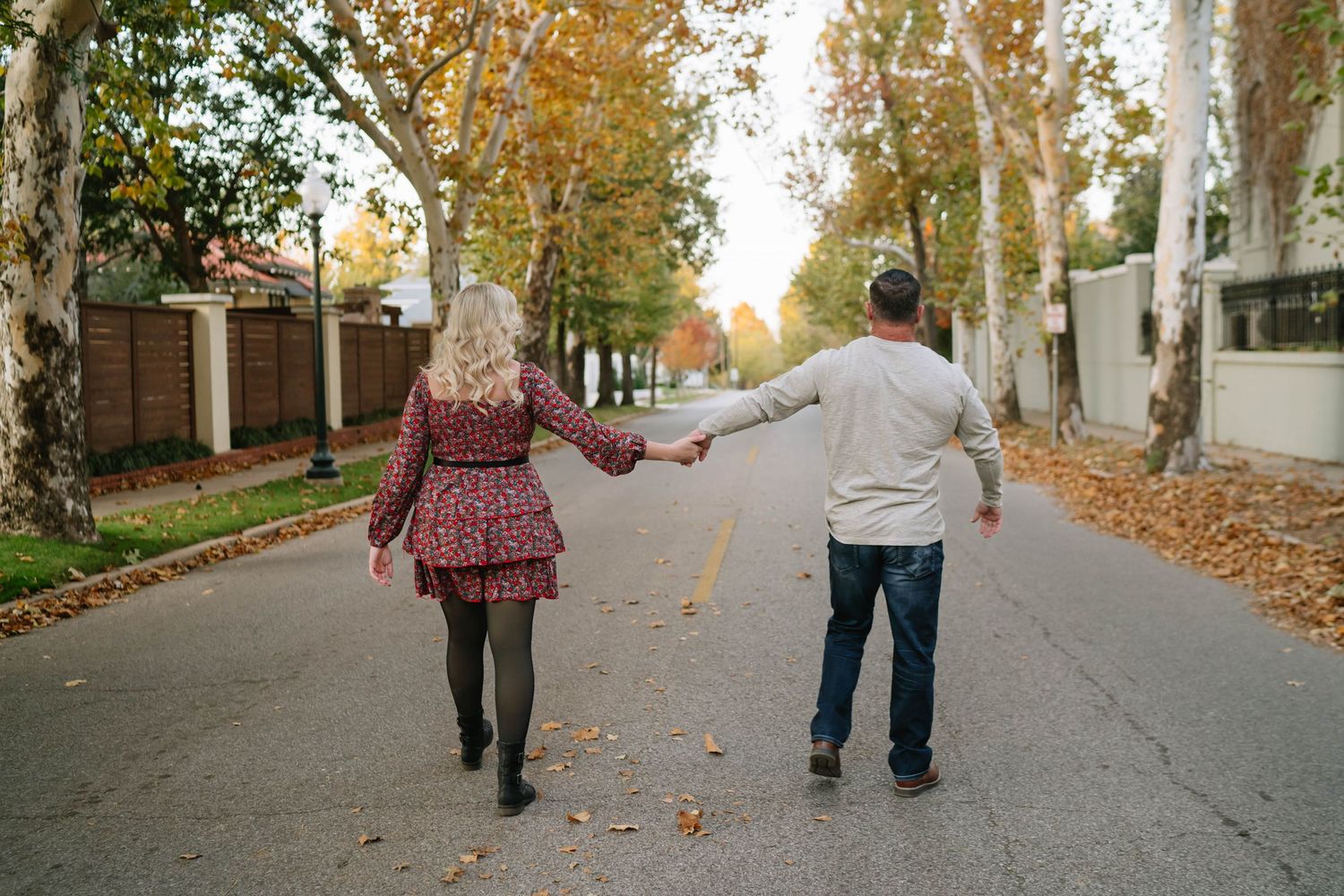 A couple walks hand in hand down a tree-lined street with autumn leaves scattered on the ground.
