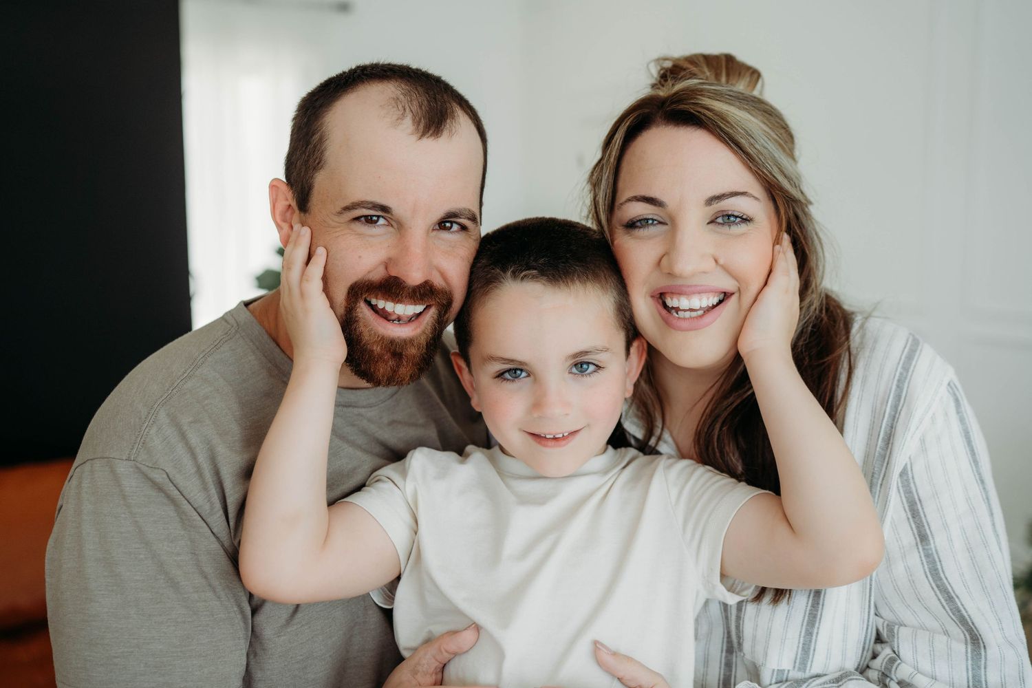 A happy family lounges together on a light gray couch wearing casual white and gray clothing for a relaxed photo session.