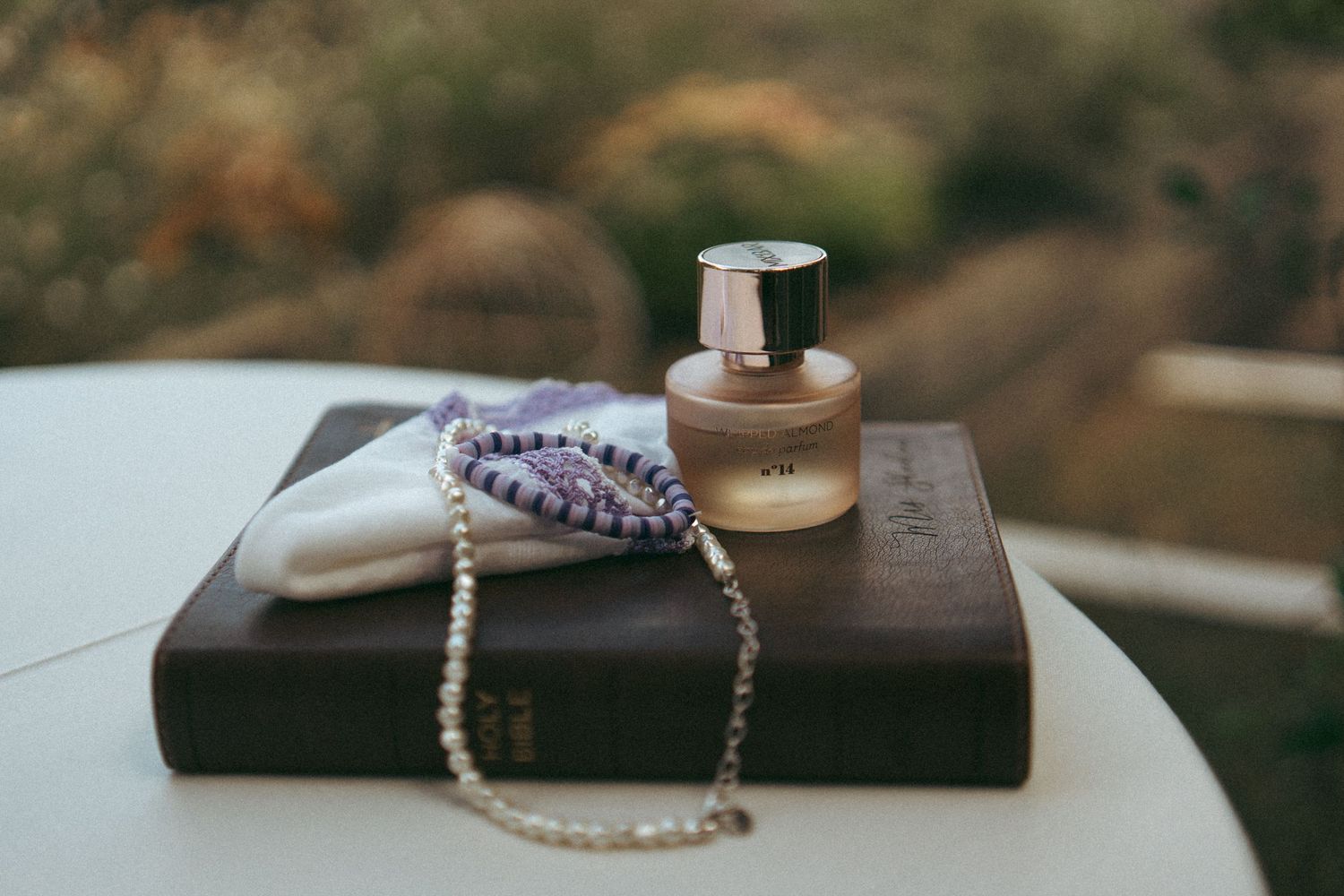 Perfume bottle and pearl necklace resting on an old book on a white table outdoors.