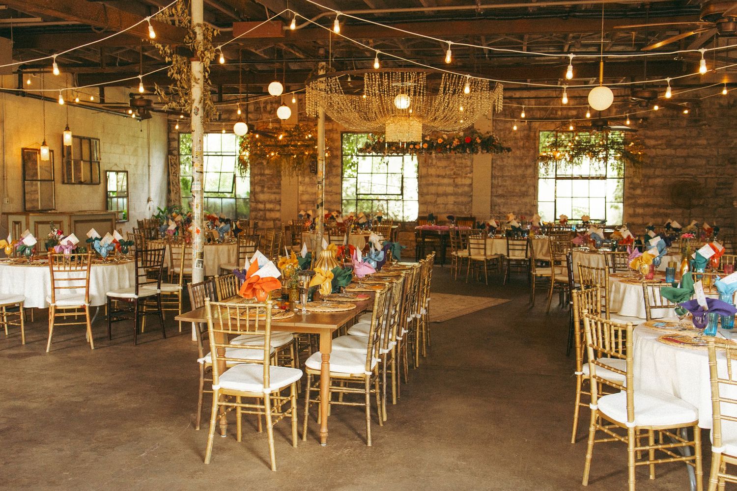 Spacious rustic wedding reception hall with gold chiavari chairs, string lights, and colorful table centerpieces.